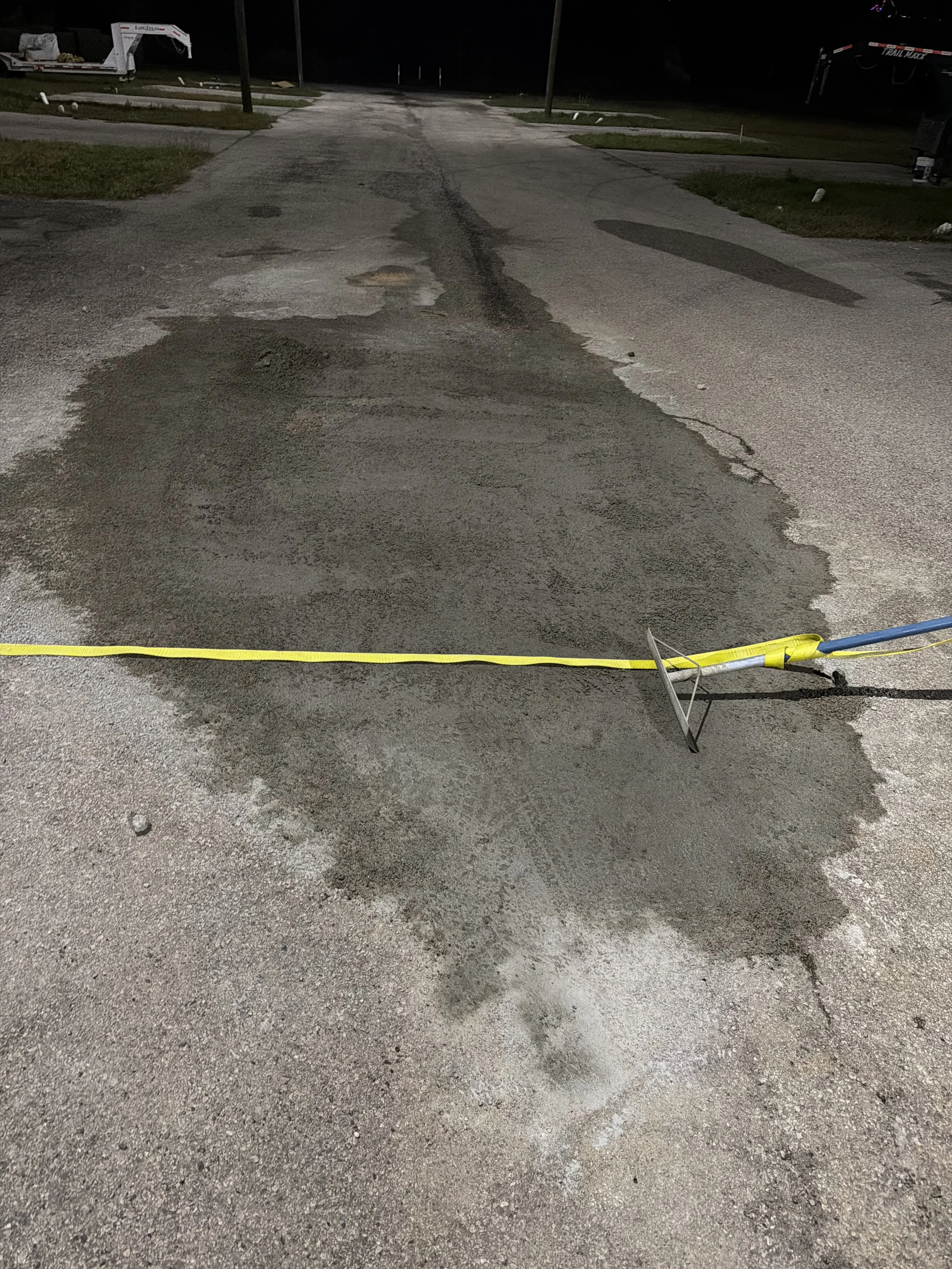A section of asphalt road under repair, with fresh concrete or patch material spread over a hole, marked off with a yellow caution tape at night.