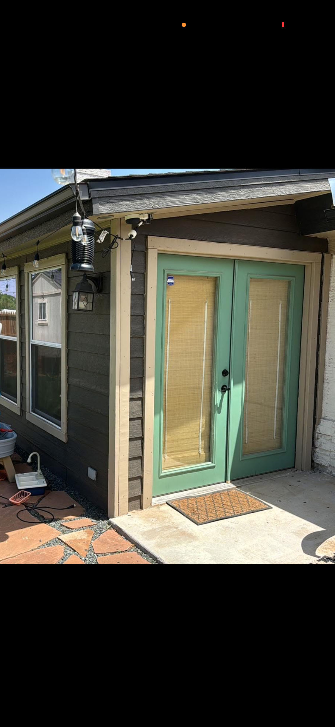 The exterior of a house with a green glass door, bamboo blinds, and a concrete patio. There are string lights and outdoor fixtures hanging from the roof.