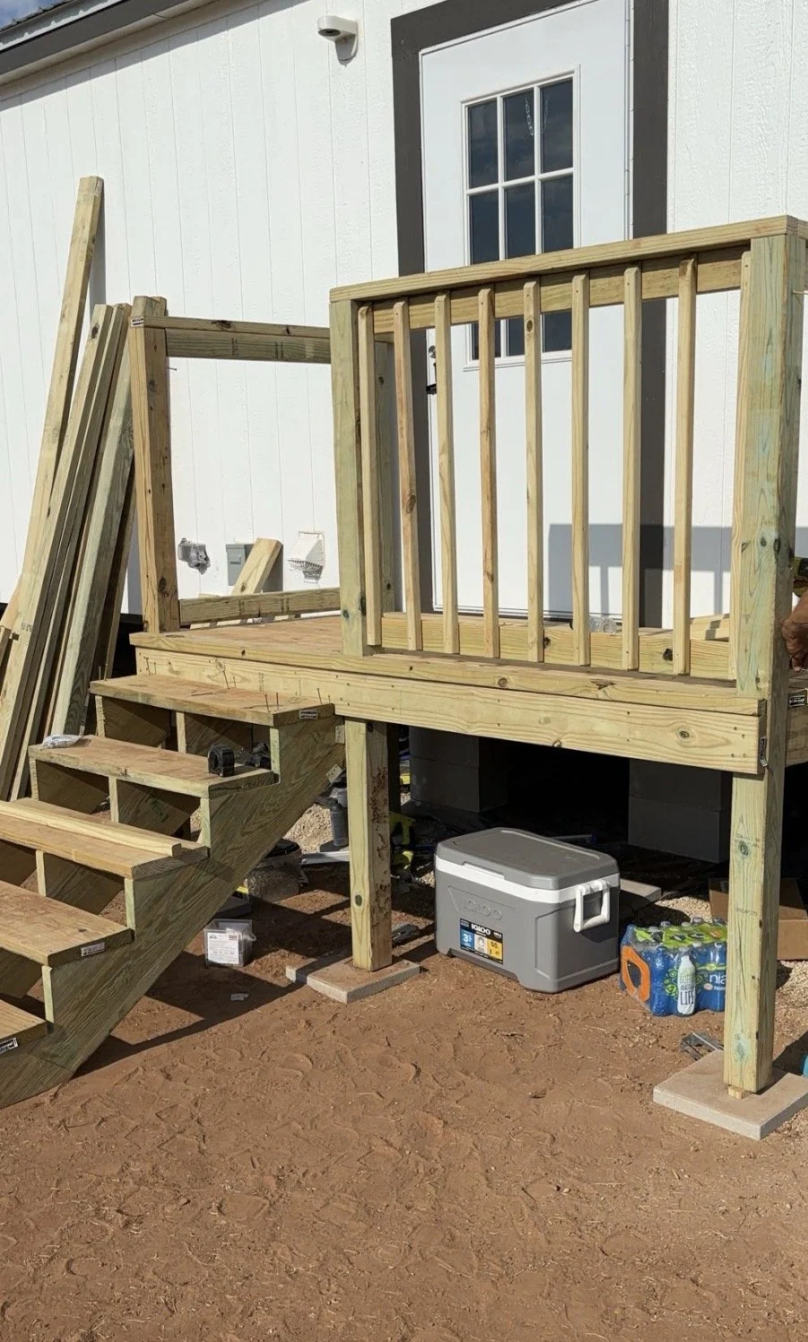 A wooden deck under construction attached to the back of a house. The deck has stairs, a railing, and a platform, with building tools and supplies nearby.