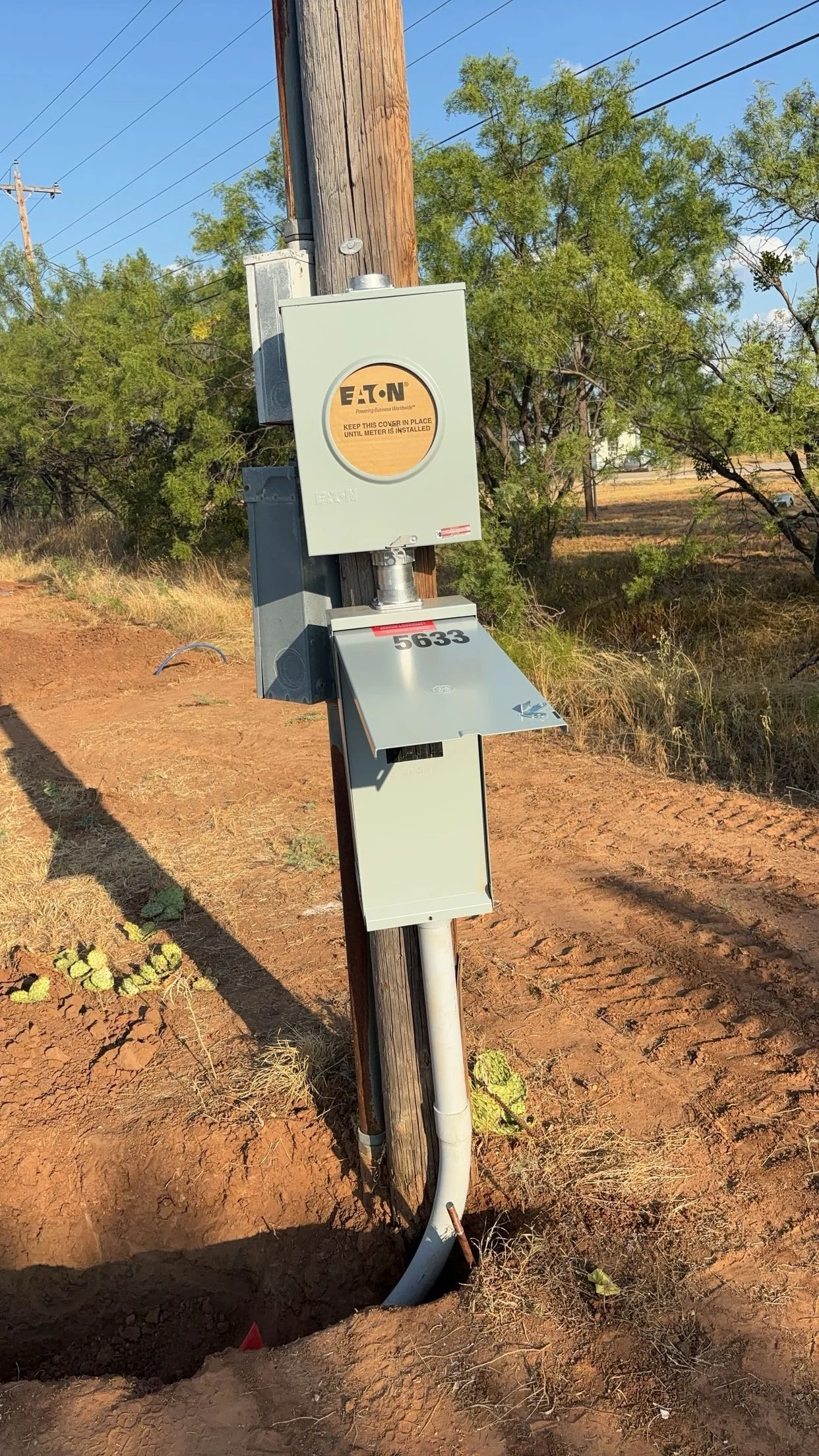 An electrical utility box mounted on a wooden utility pole with a metal cover in an outdoor setting. The background includes trees, power lines, and an unpaved dirt ground with some cactus plants.