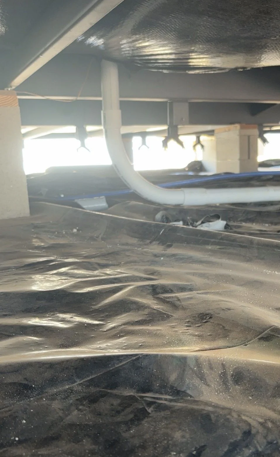 Under a parking structure with visible pipes and support beams, a dusty, uneven floor surface is shown with a black tarp or covering.