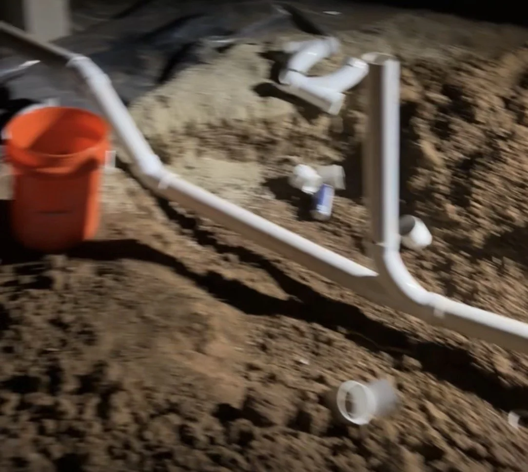 Construction site with exposed white PVC pipes, an orange bucket, and dirt ground.