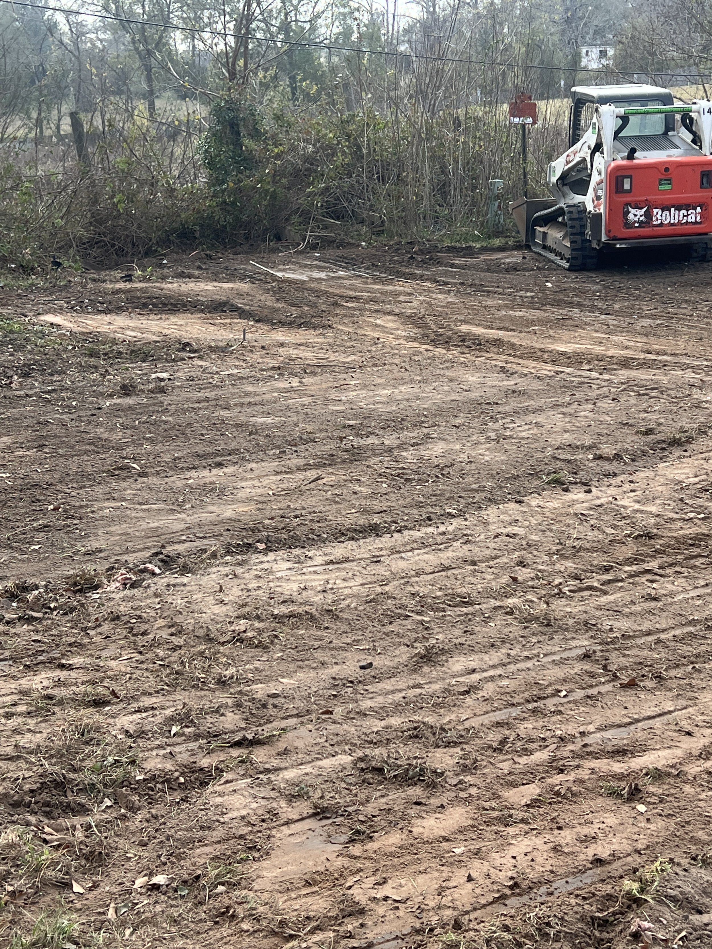 A cleared dirt area with tire tracks, adjacent to a wooded area with bushes and trees, and a small Bobcat construction vehicle parked on the right side.