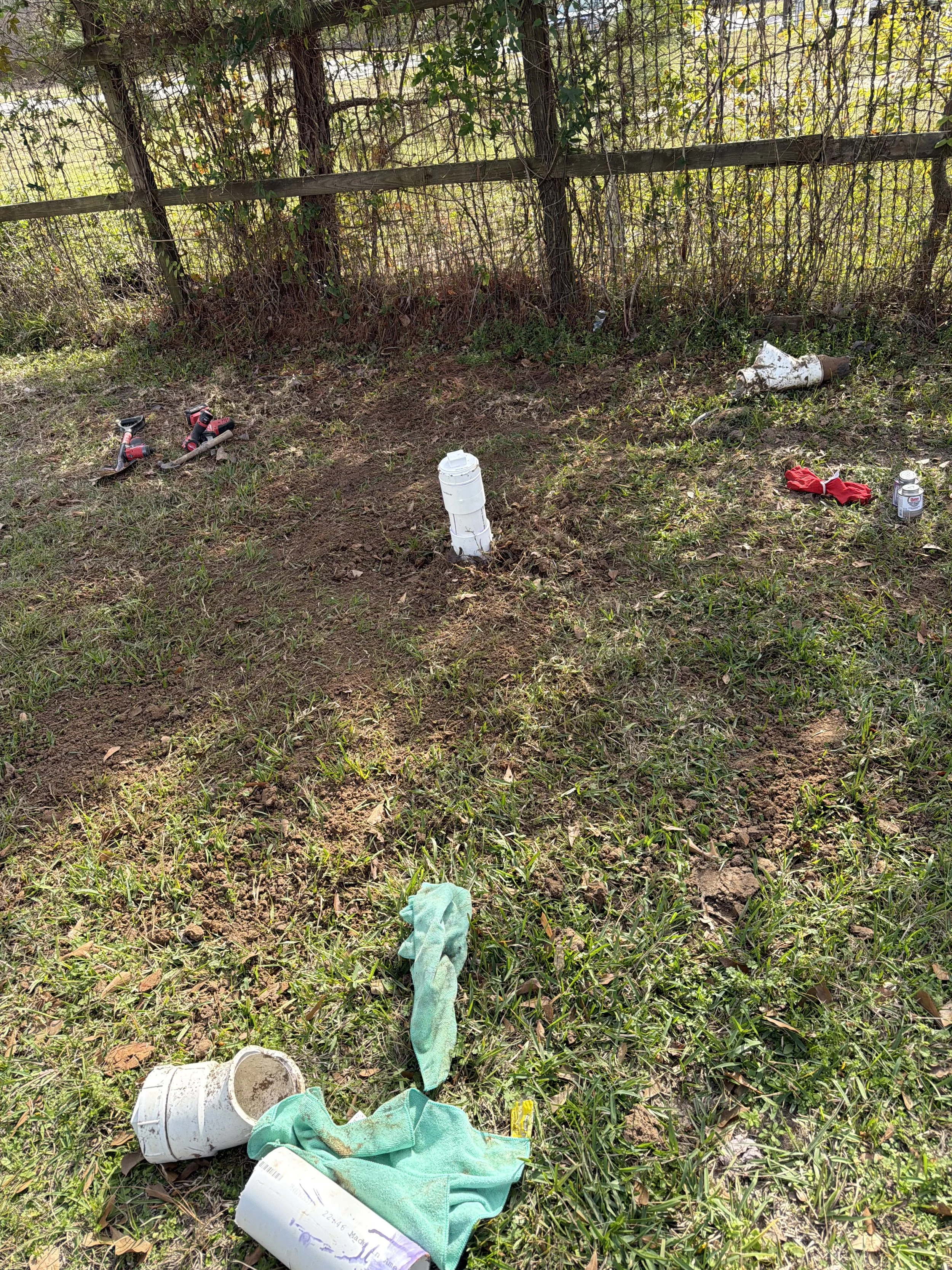 Littered yard with upside-down trash can, discarded cups, gloves, and cans near a wooden fence with overgrown bushes.