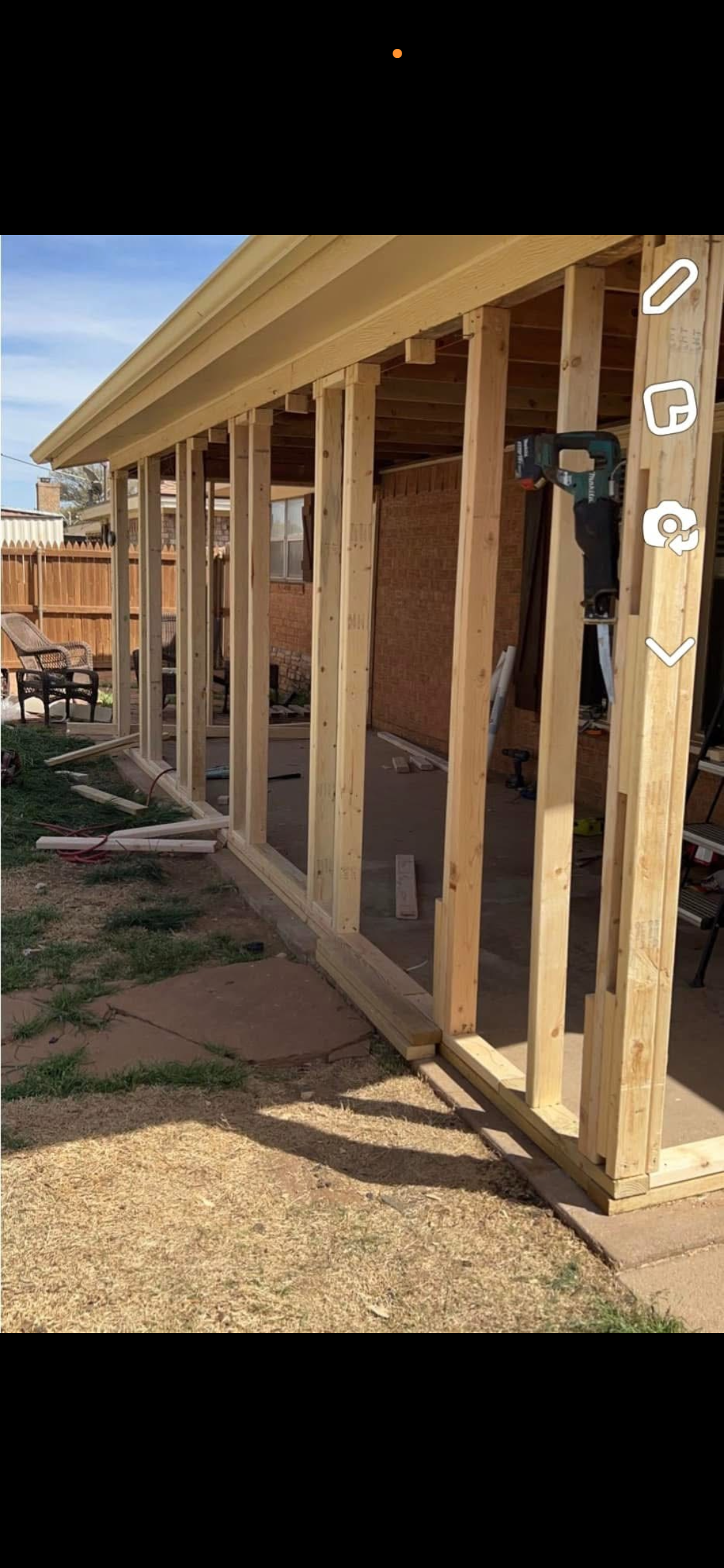Wooden framing of a small outdoor structure in a backyard, with a wooden fence and patio chairs in the background.