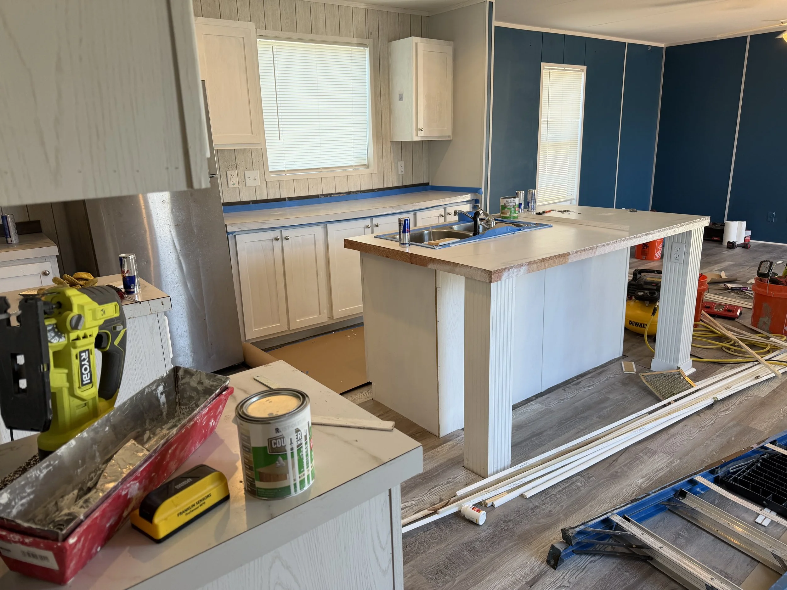 Kitchen under construction with tools and materials scattered around, including a cordless drill, paint cans, molding, and partially installed cabinetry.