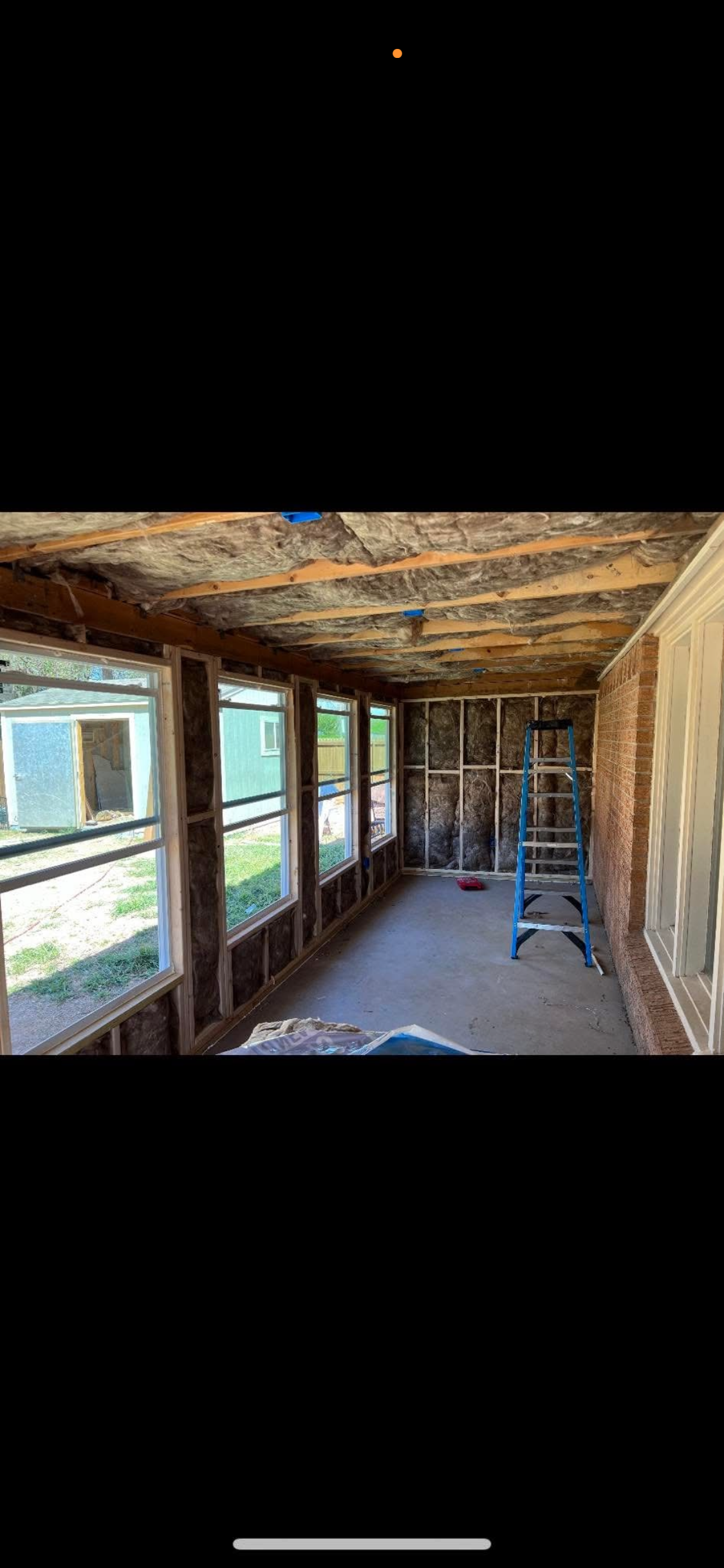 Interior of a room under renovation with exposed insulation, wooden studs, and a stepladder, windows along one wall, and construction tools on the floor.