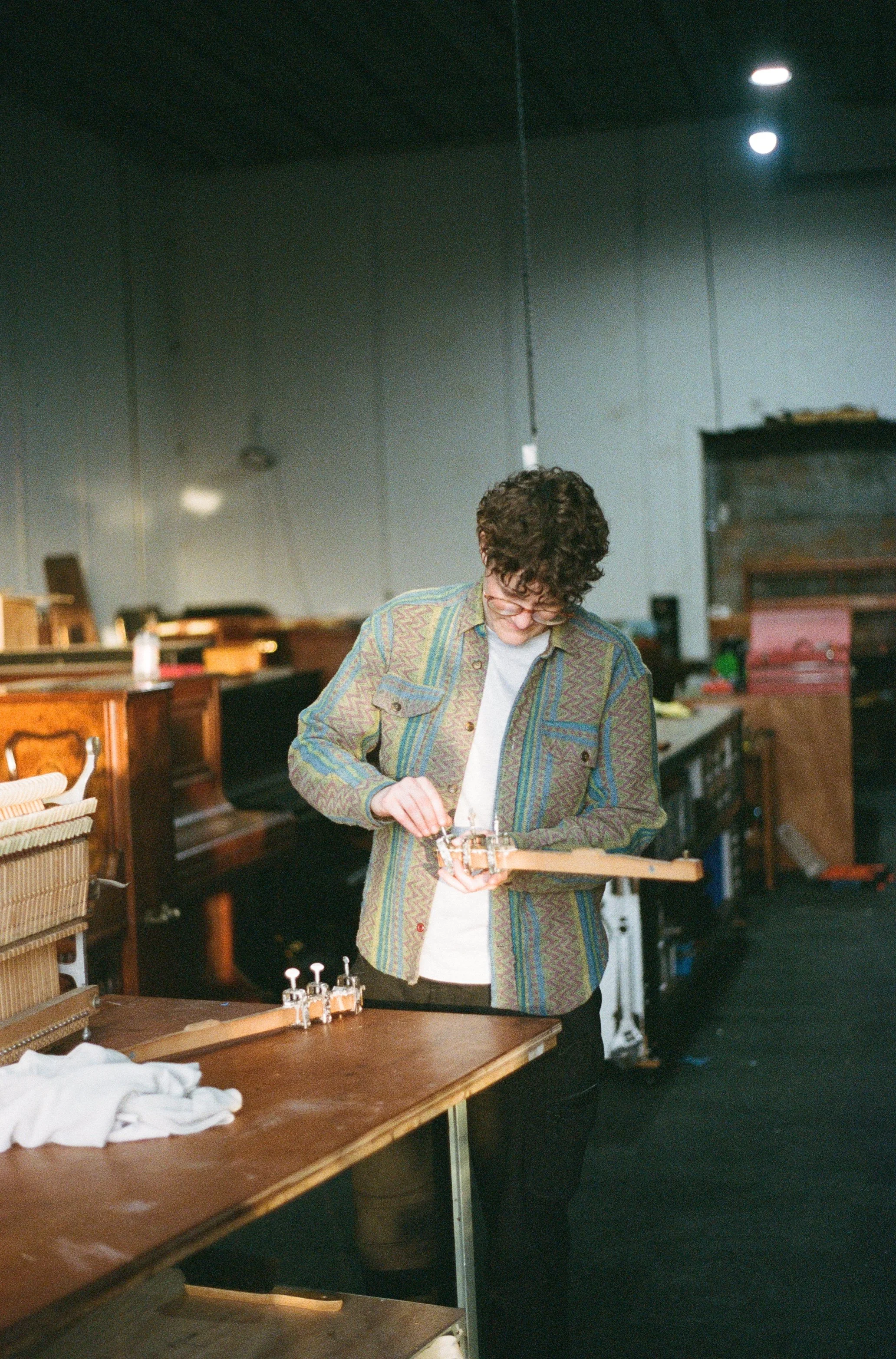 A person with curly hair and glasses working on a small woodworking project in a room filled with furniture and tools.
