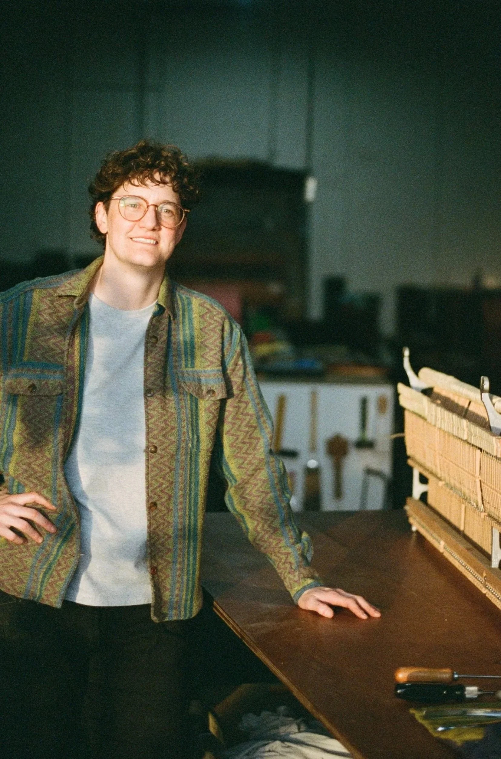 A person with short curly hair, glasses, wearing a patterned jacket, standing in a workshop or studio, with tools on a table and a dark background.