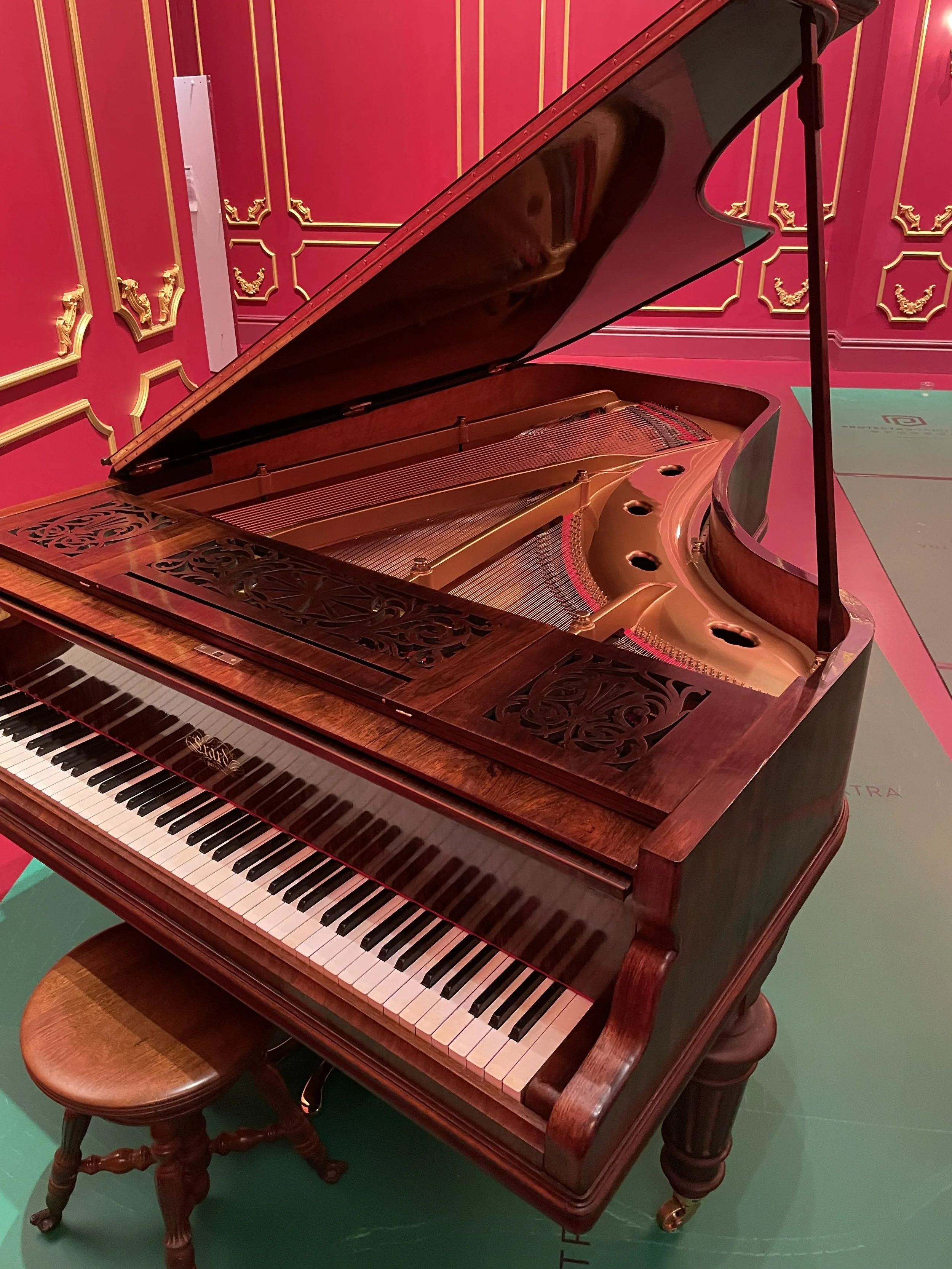 A grand piano with a polished wooden finish, open lid, and black and white keys, situated on a green floor against a decorative pink wall with gold accents.