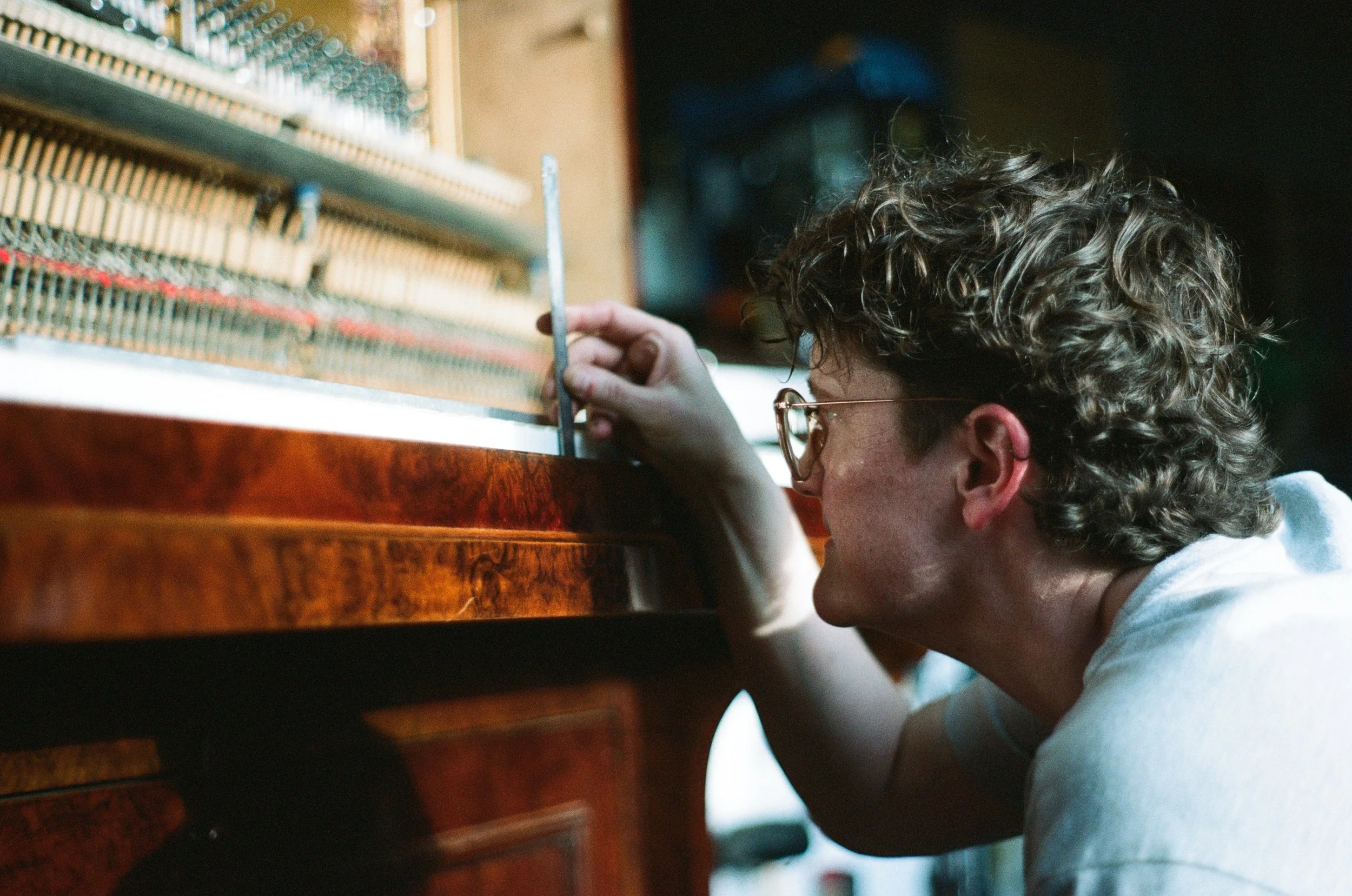 Person with curly hair and glasses working on piano strings with a tuning tool.