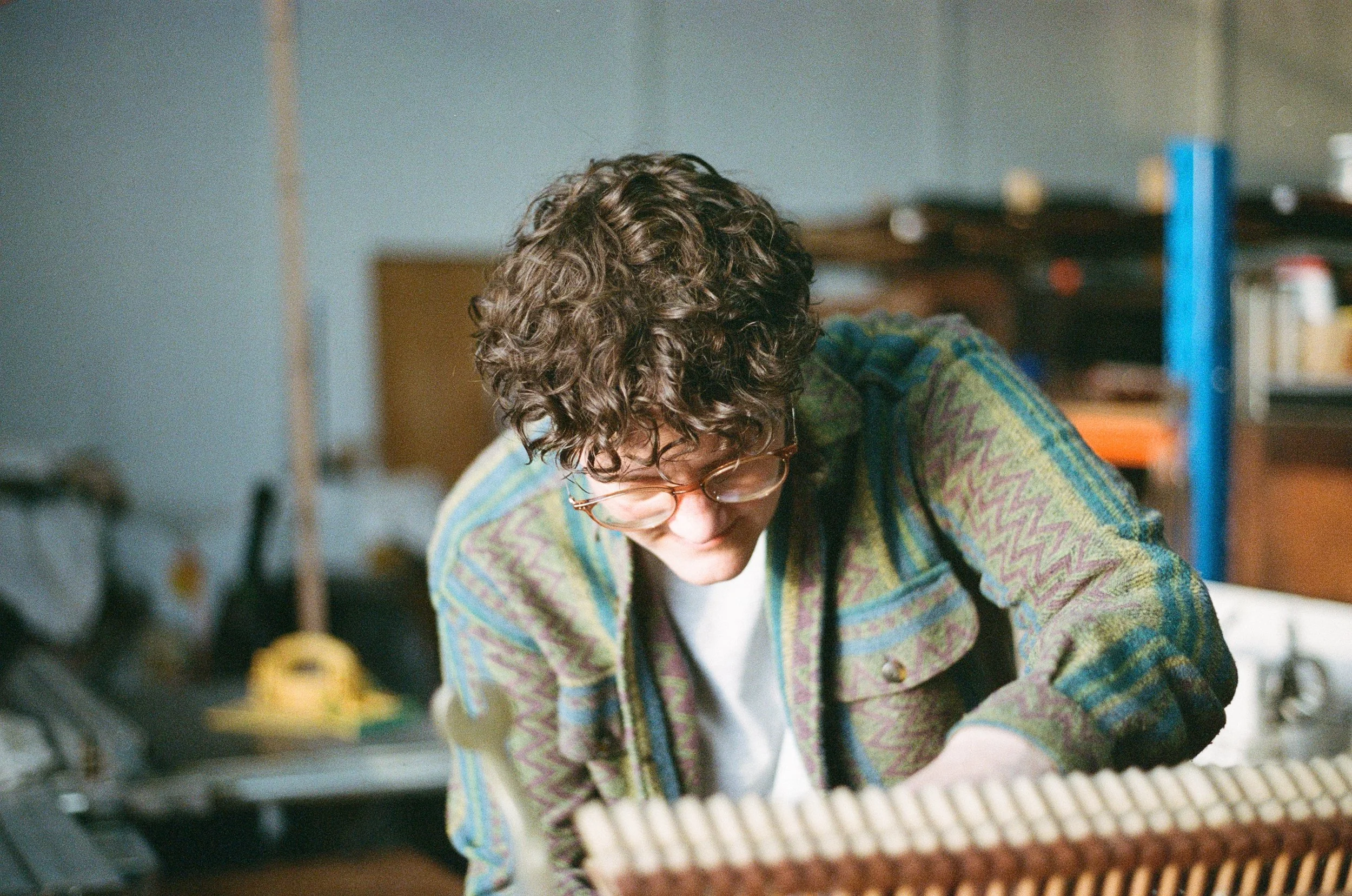 A person with curly hair and glasses wearing a colorful, patterned jacket working on a project at a cluttered workbench in a workshop or garage.