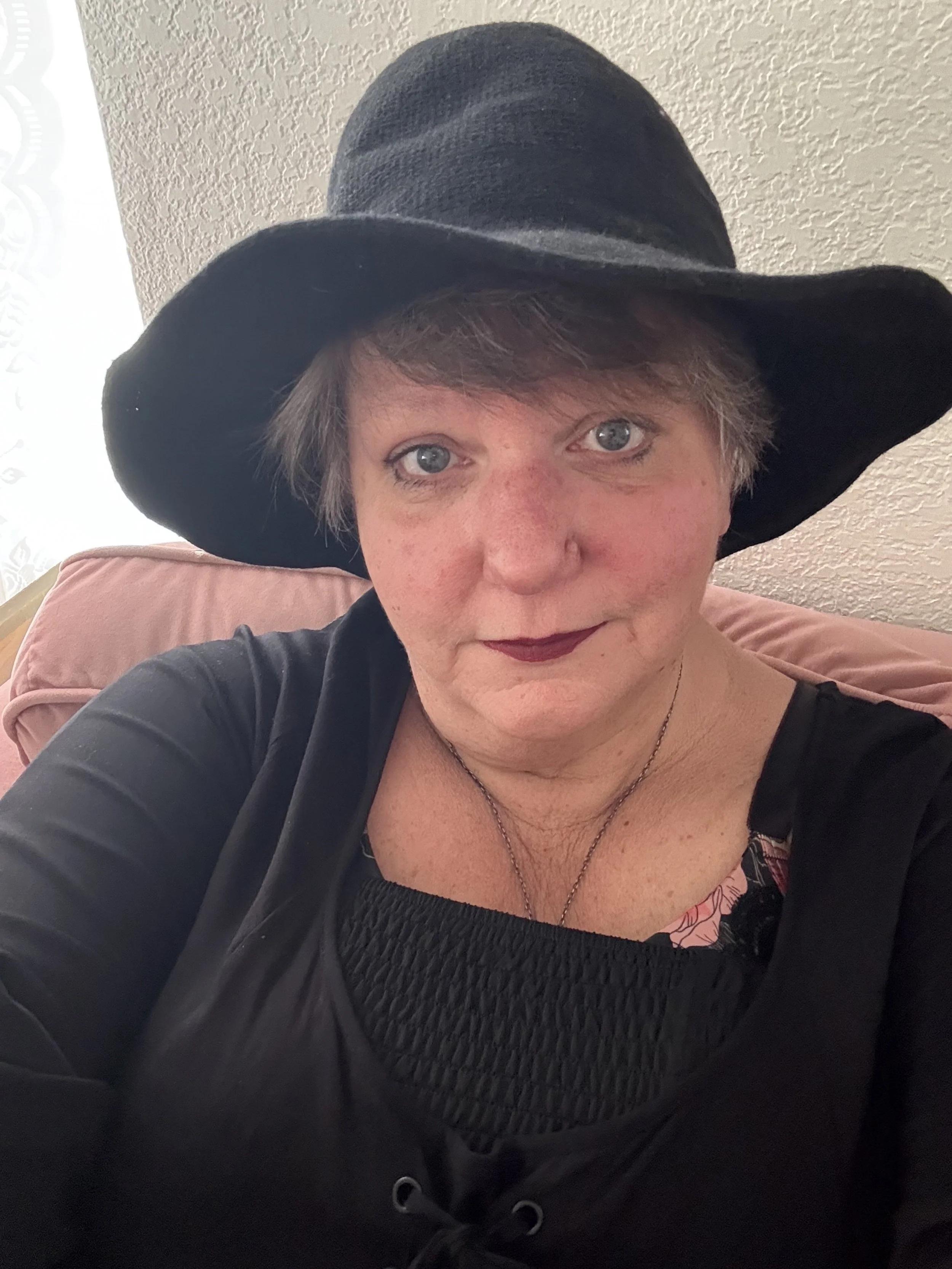 A woman wearing a large black floppy hat, black top with textured fabric, and dark lipstick, sitting on a pink cushion in a room with textured beige walls.