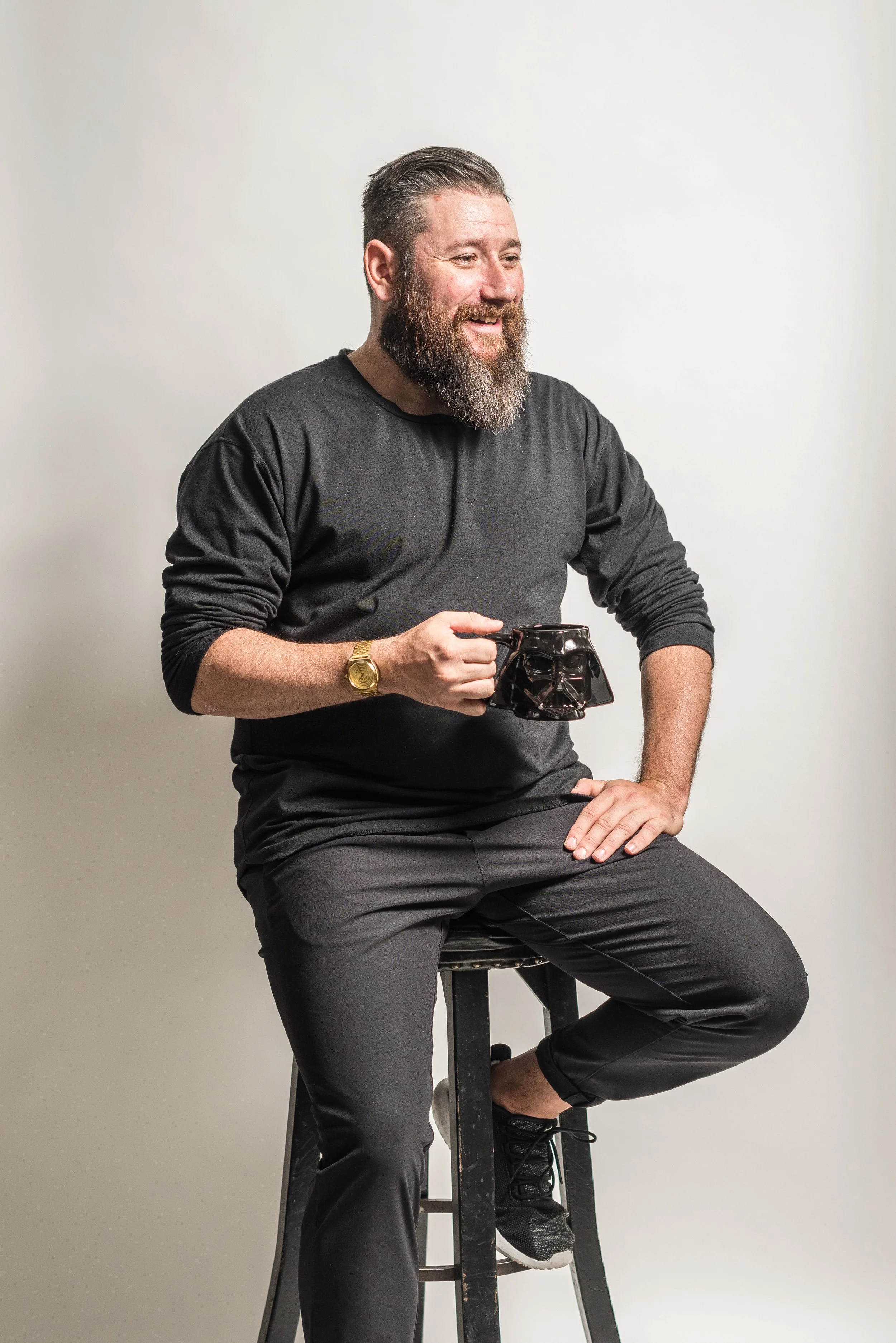 A man with a beard sitting on a stool, holding a Darth Vader mug, smiling, dressed in black, against a plain white background.