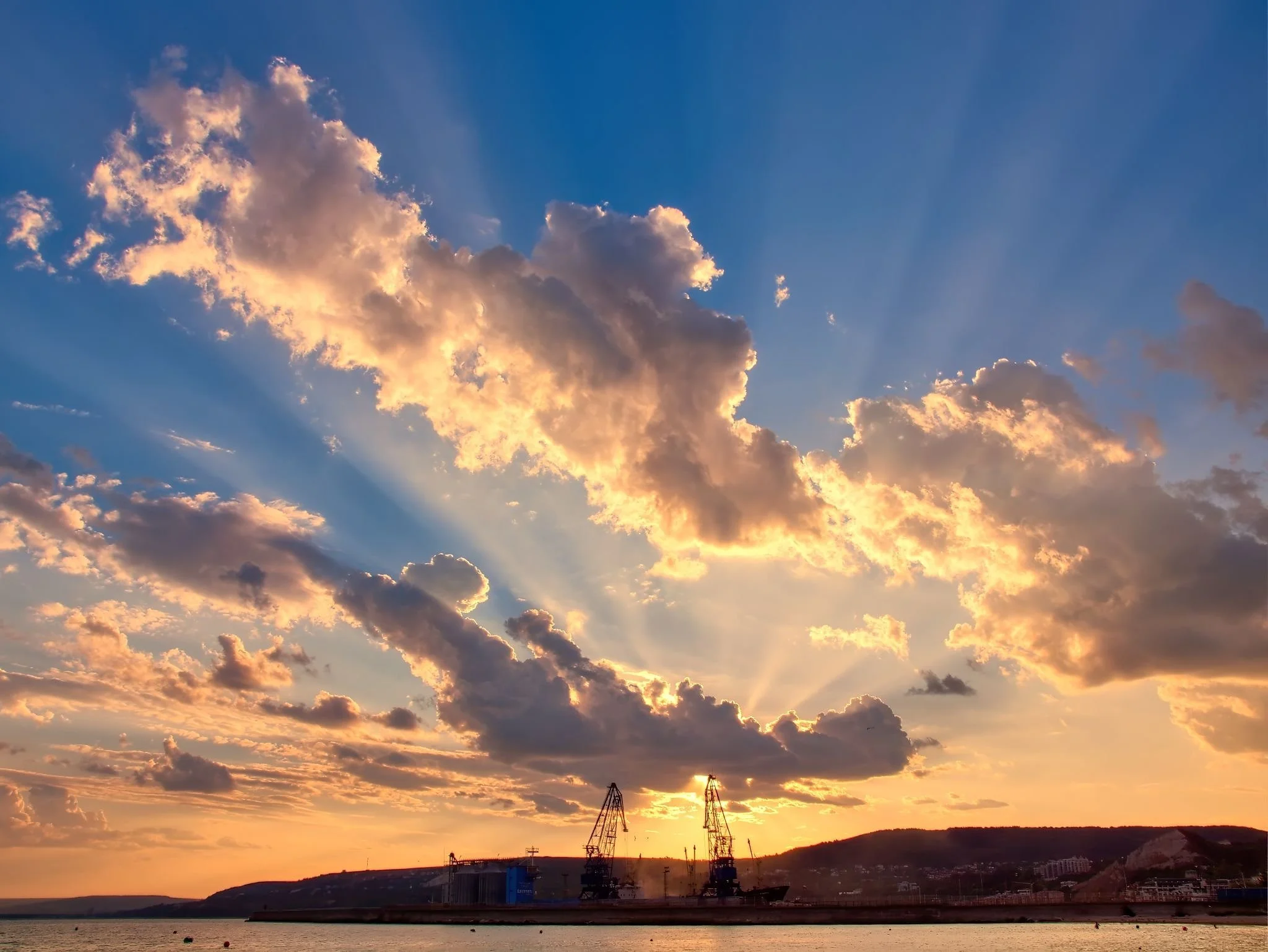 Auringonlasku Balchikin satamassa Bulgariassa, jossa nosturien siluetit näkyvät pilvisellä taivaalla Mustanmeren rannalla.
