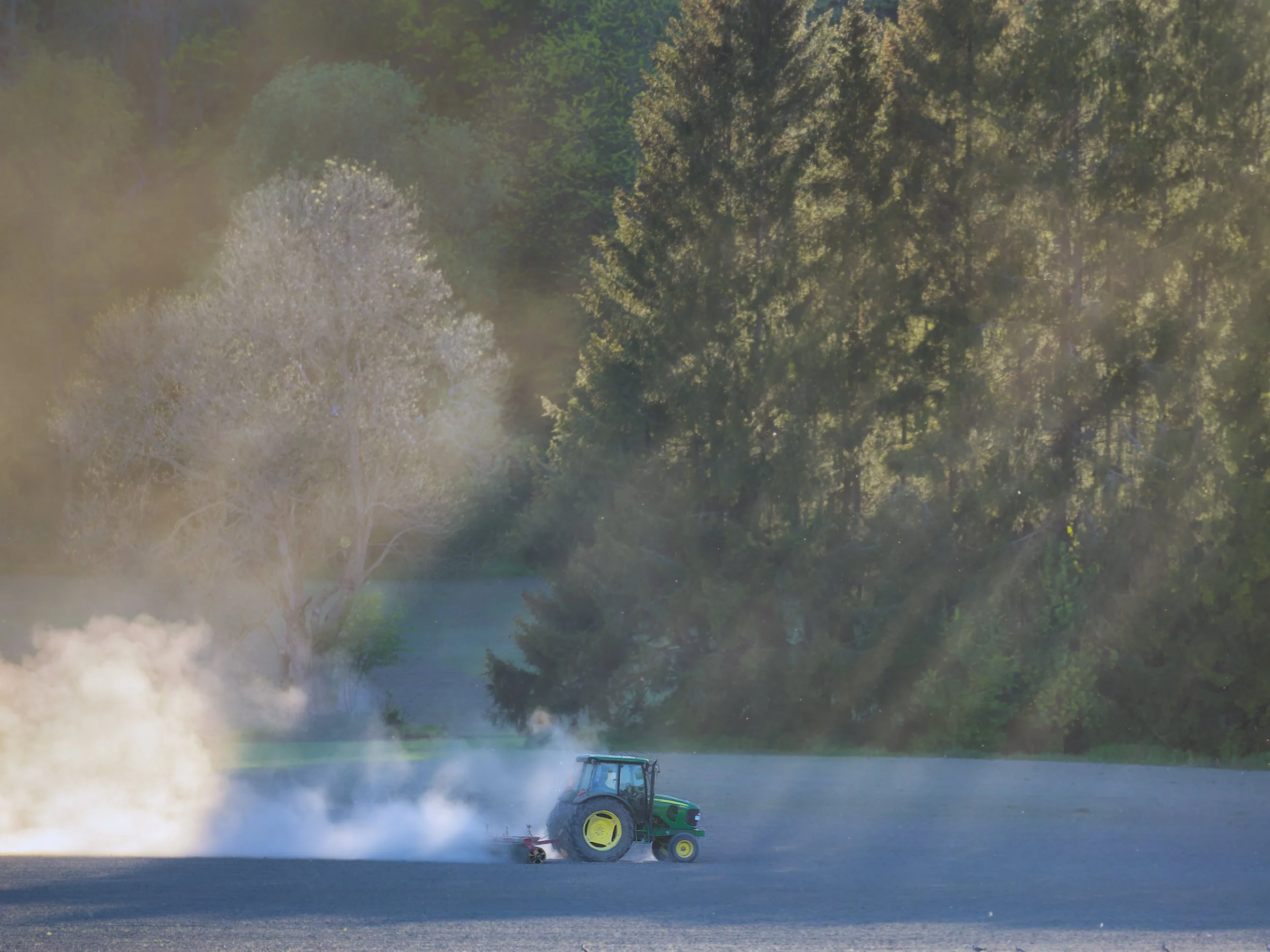 Tunnelmallinen valokuva maataloustöistä, jossa John Deere traktori muokkaa kuivaa peltoa vastavalossa. Kuva korostaa suomalaista maaseutua ja kevään kiireisiä viljelytöitä.