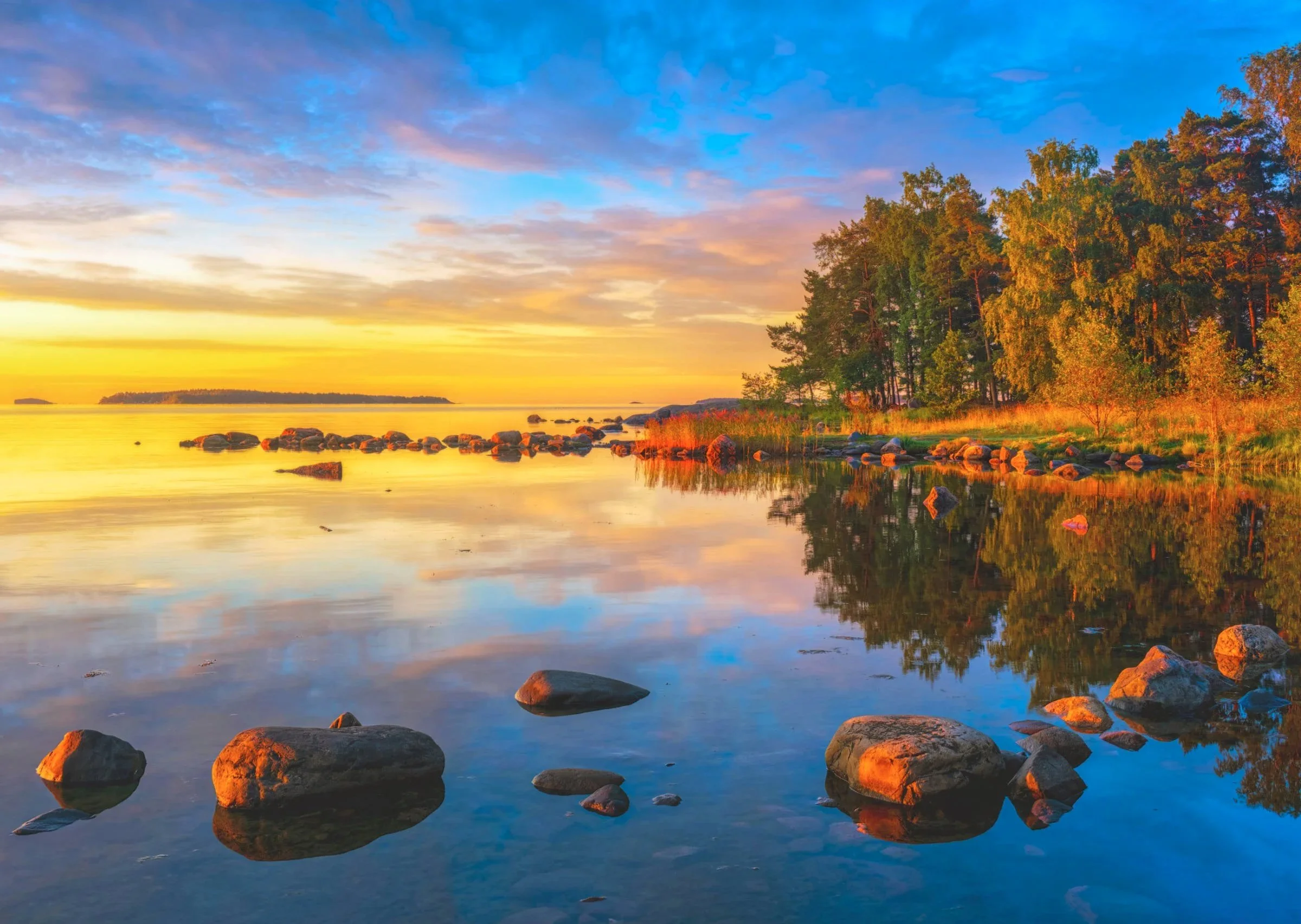 Hehkuvat auringonnousun värit maalaavat tyynen Itämeren Uutelassa Helsingissä; hohtavat kivet ja puustoinen rantaviiva heijastuvat peilityynestä vedestä.