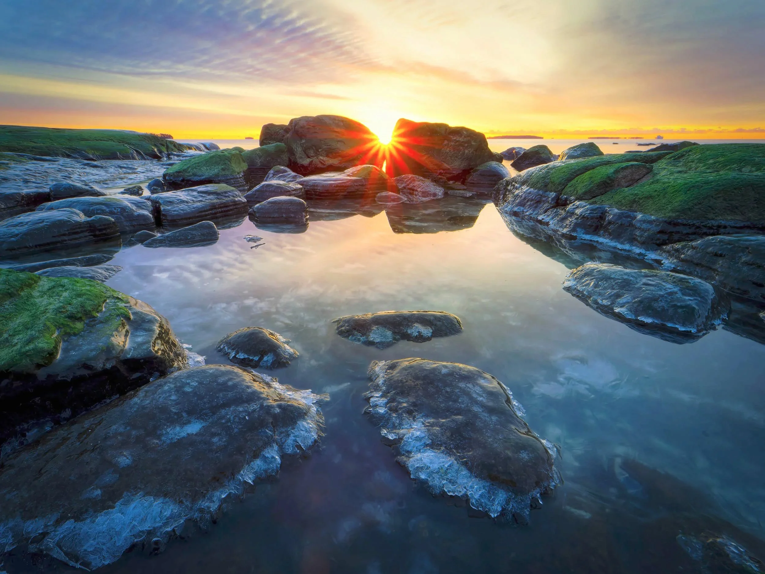 Kylmä auringonnousu Uutelassa Helsingissä, aurinko piirtää kirkkaan tähden sammaleisten, jäärreunaisten kivien ylle, ja tyynet rantavesialtaat heijastavat pastellinsävyistä Itämeren taivasta.