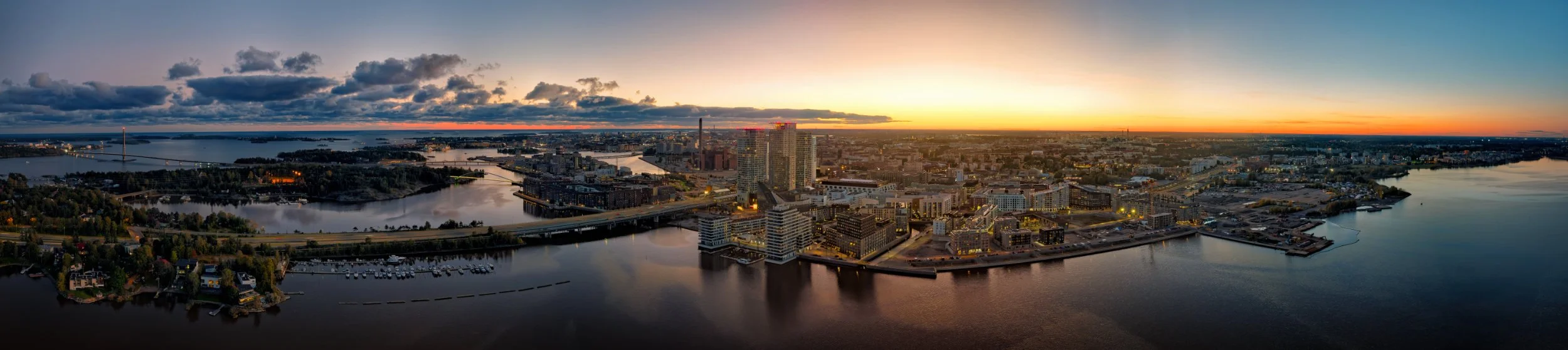 Panoraama-ilmakuva Helsingin Kalasatamasta auringonlaskun aikaan. Kuvassa näkyvät Kalasataman pilvenpiirtäjät (Majakka ja Loisto), Kulosaaren silta sekä iltahämärässä valaistu kaupunkisiluetti meren rannalla.