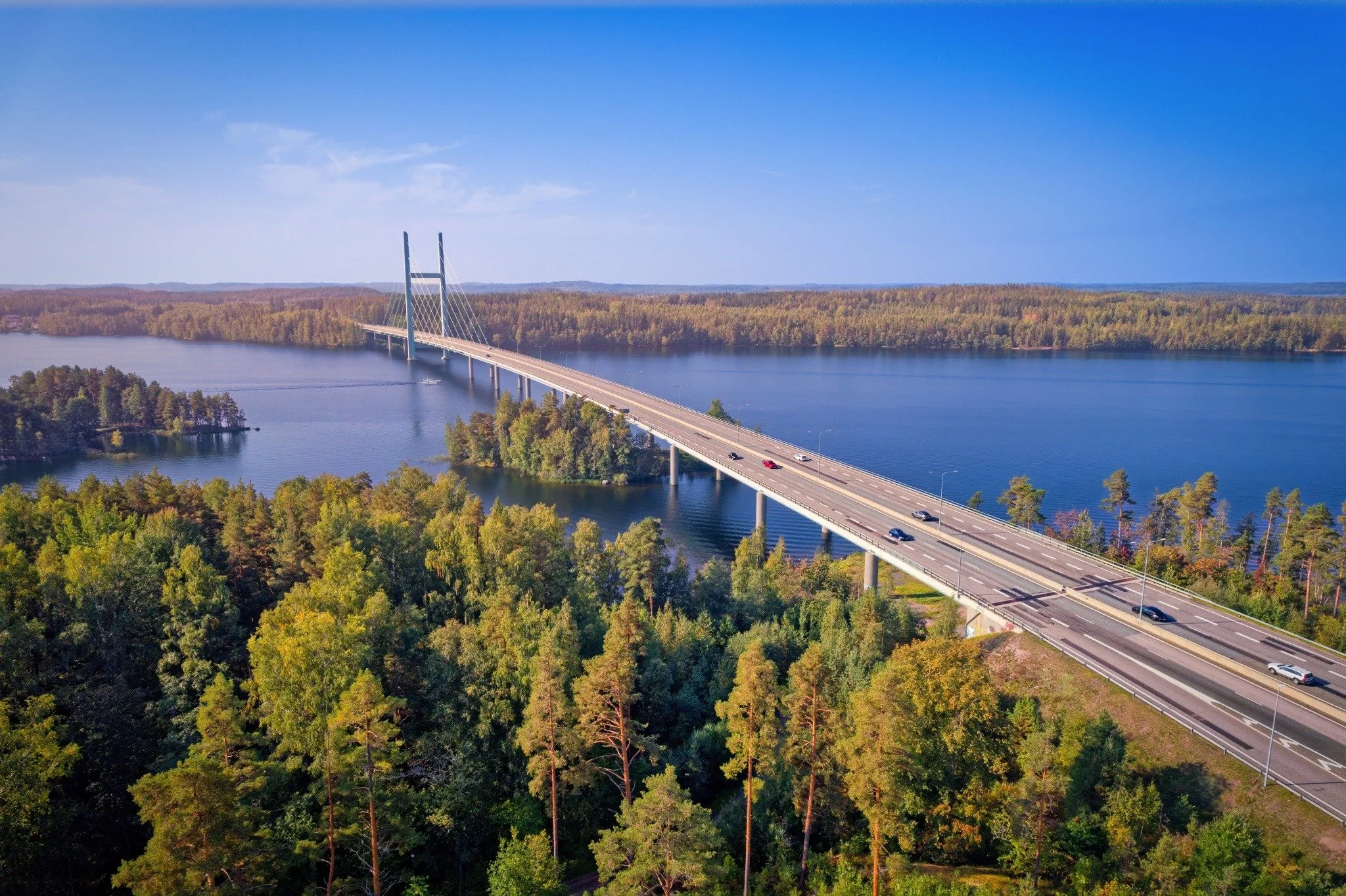 Majesteettinen ilmakuva Tähtiniemen sillasta Heinolassa. Tämä vaikuttava vinoköysisilta ylittää kimaltelevan sinisen järven ja yhdistää rehevät vihreät metsäsaaret ja mantereen. Näkymä kylpee kirkkaassa auringonvalossa selkeän sinisen taivaan alla, j