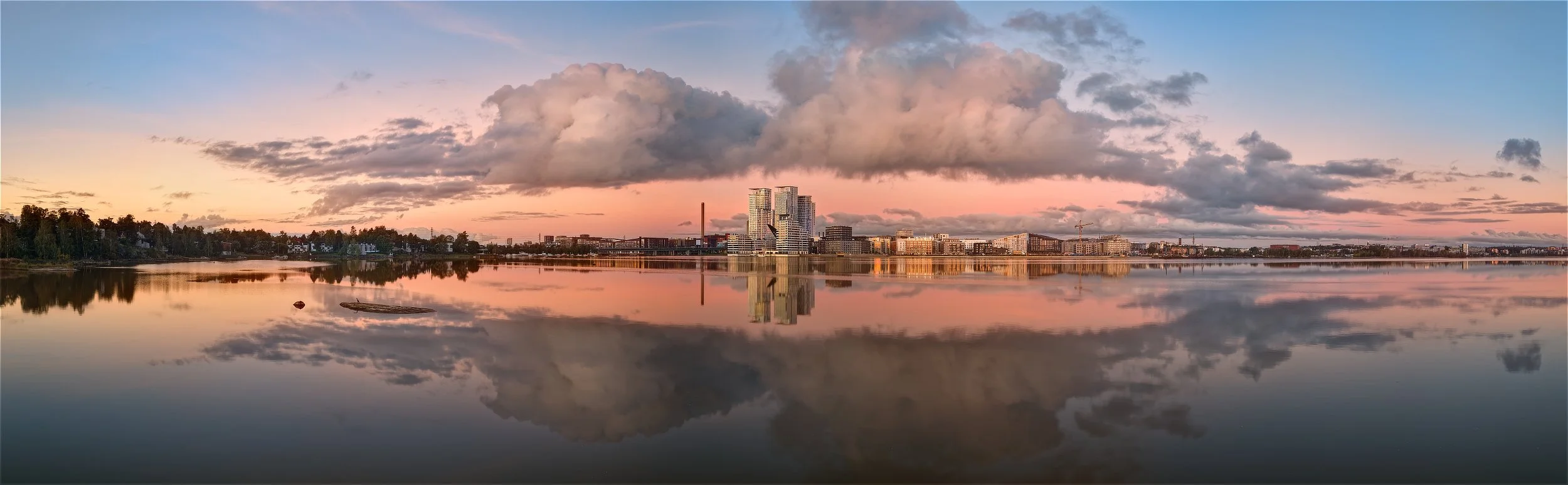Ilmakuva DJI-dronella kuvatusta panoraamasta Kalasatamassa Helsingissä auringonnousun aikaan; pastellinsävyiset pilvet ja modernit rantarakennukset heijastuvat täydellisesti tyynen lahden pinnasta.