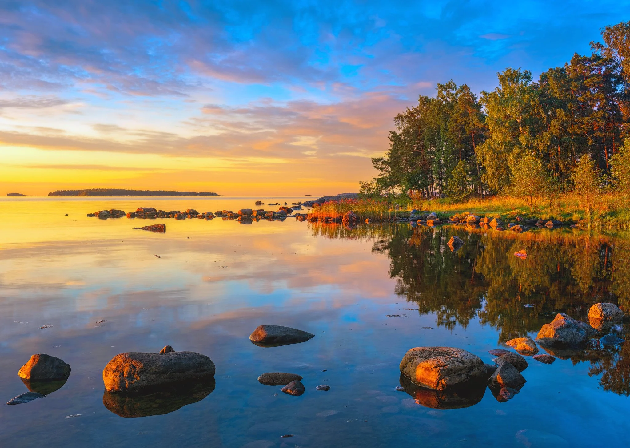 A glassy summer morning at Uutela, Helsinki. Warm golden sunrise light 
bathes the granite boulders and reed beds along the shore, while pine 
and birch trees glow against a vivid blue and orange sky. The still 
water mirrors the scene almost perfect