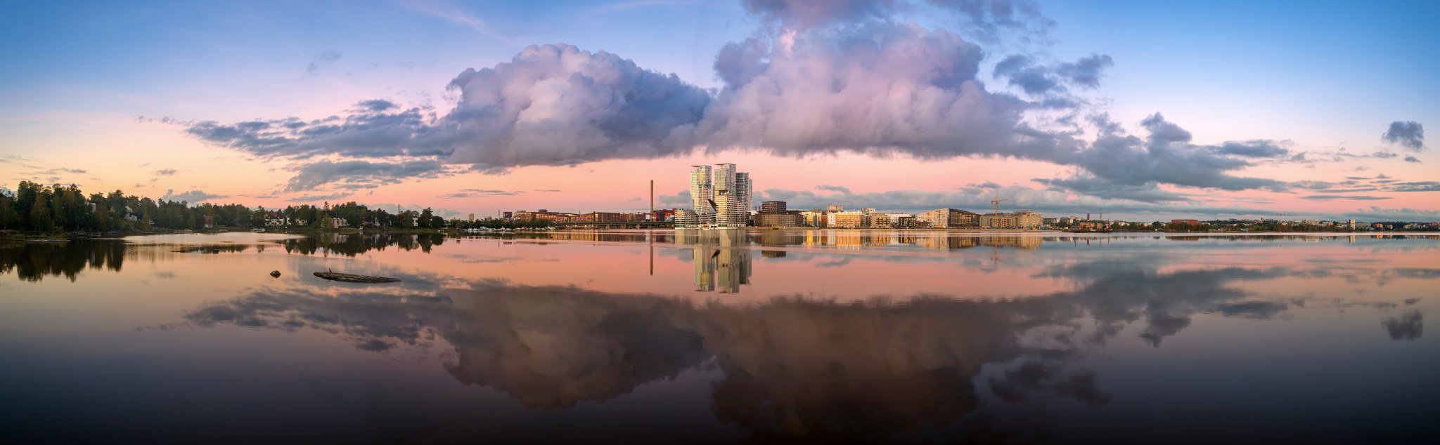 Ilmakuva DJI-dronella kuvatusta panoraamasta Kalasatamassa Helsingissä auringonnousun aikaan; pastellinsävyiset pilvet ja modernit rantarakennukset heijastuvat täydellisesti tyynen lahden pinnasta.
