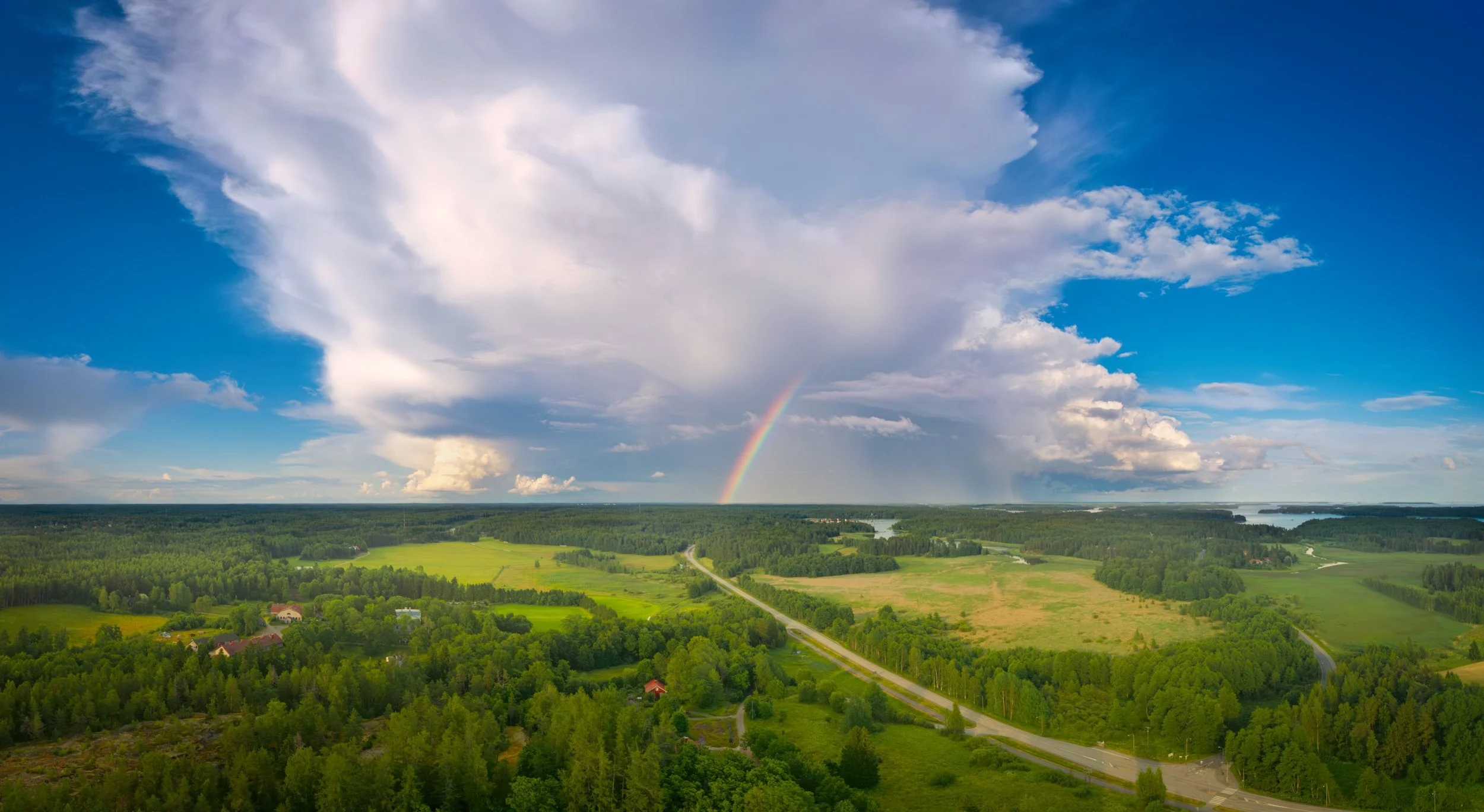 A breathtaking wide panoramic aerial view of a lush green summer landscape under a dramatic blue sky. A massive storm cloud dominates the sky, with a vibrant rainbow stretching down to the dense forests, fields, and a winding road below.