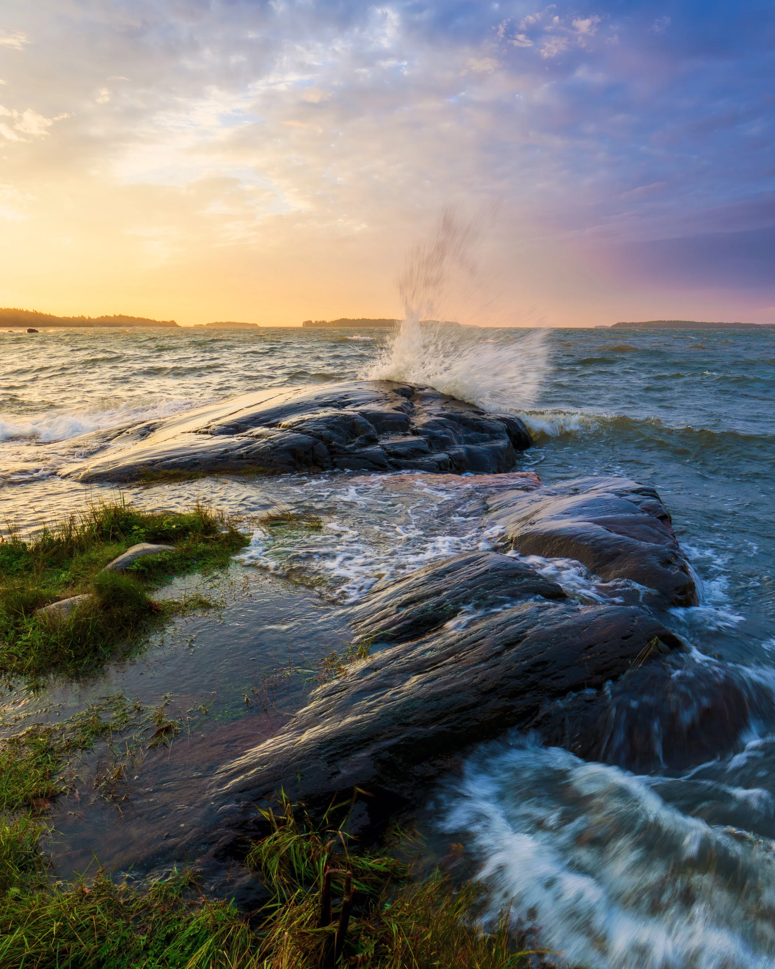 Kultainen auringonlaskun valo osuu Itämereen, kun aalto murtuu tummia kalliota vasten Uutelassa Helsingissä; etualalla hehkuvat ruoho ja pienet rantavesialtaat.