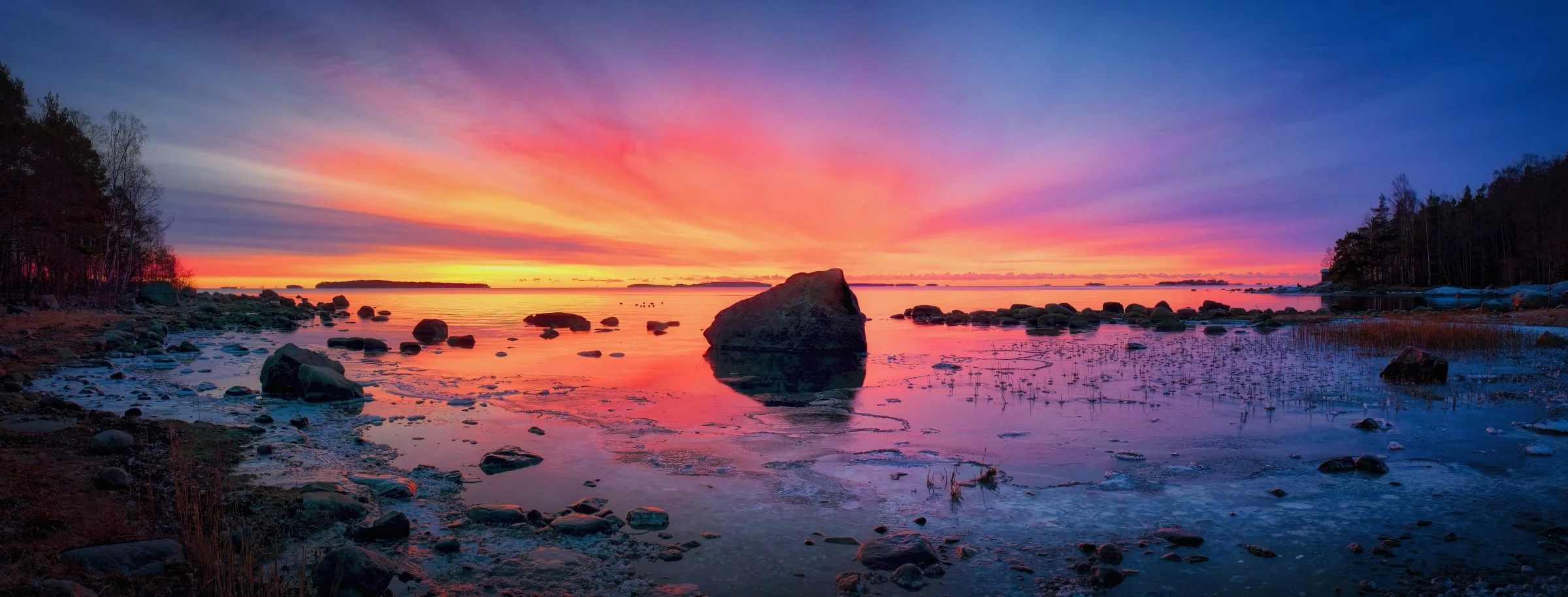 A dramatic winter sunrise panorama from Kallahti shore in Helsinki, Finland. Intense crimson and violet sky radiates above the calm Baltic Sea, reflecting warm tones across the partially frozen shoreline. Scattered boulders and reed stems emerge from