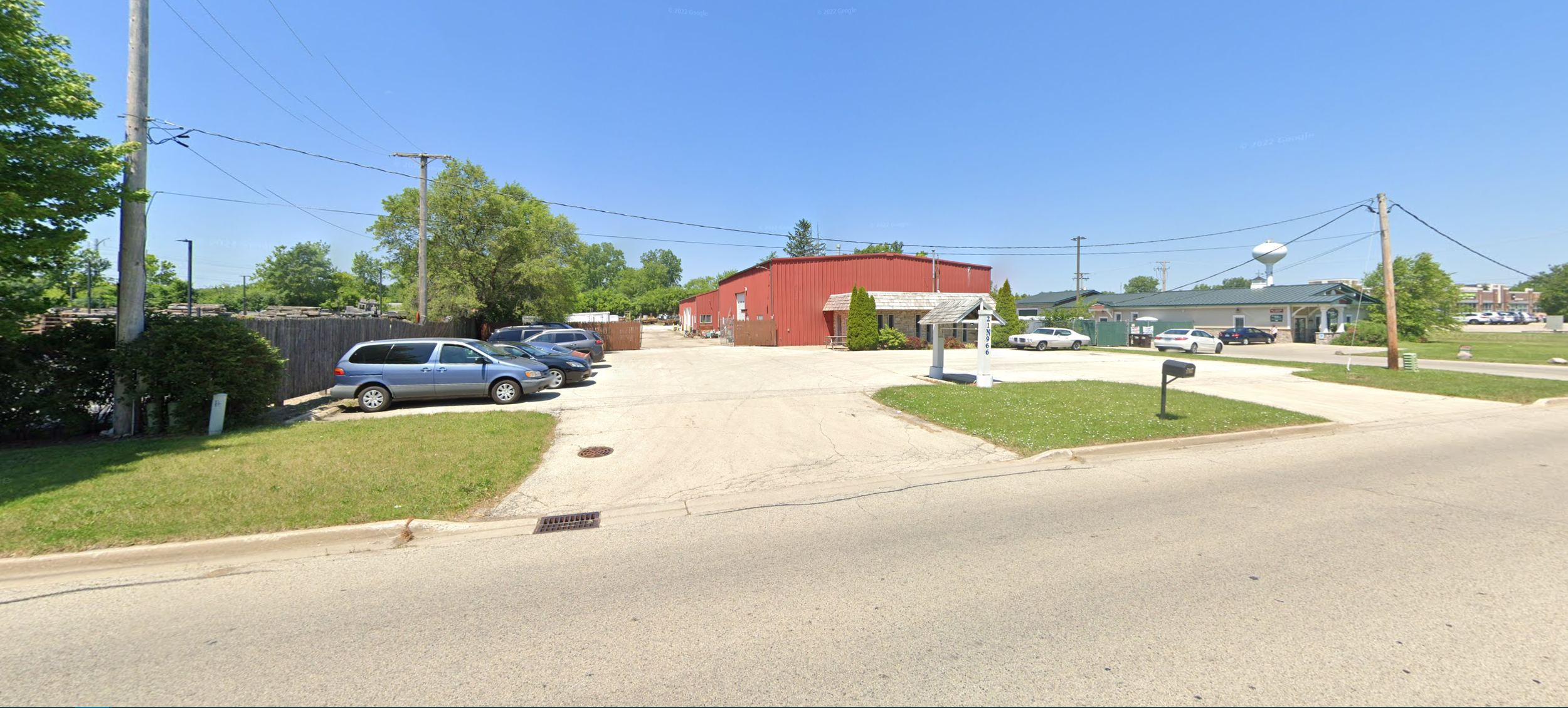 A parking lot with several cars parked near a red building and a small white structure with a sign, under a clear blue sky, with trees and power lines in the background.