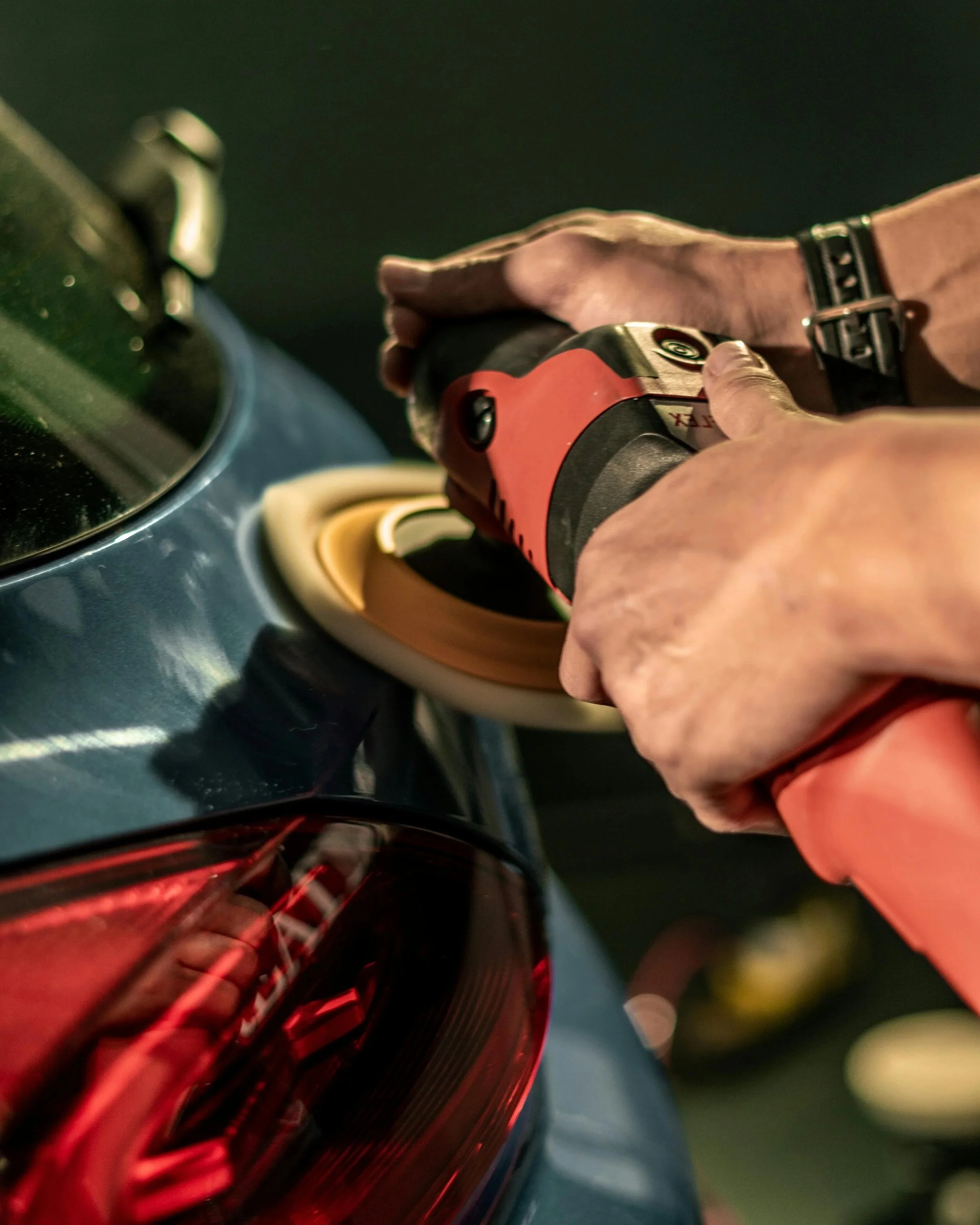 Person polishing the rear tail light of a dark-colored vehicle with a handheld polishing tool.
