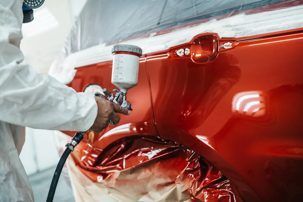 A person in white protective clothing using a spray gun to apply red paint on a car's body in an auto body workshop.