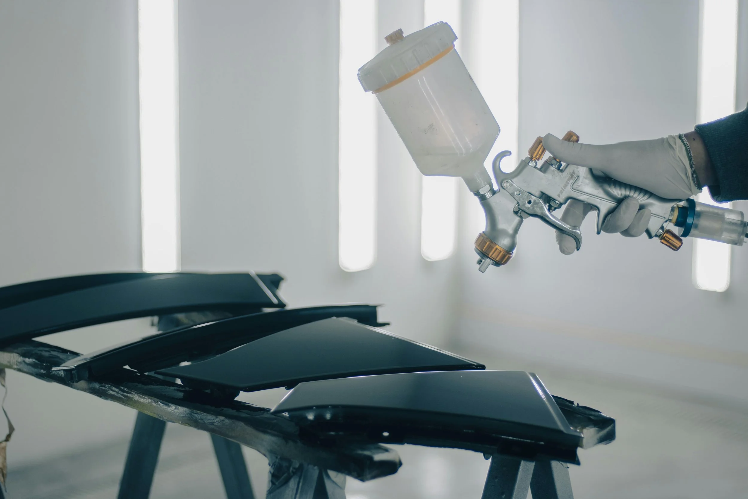 A hand wearing a glove holding a spray gun, used for painting, with black painted surfaces on stands in a well-lit room.