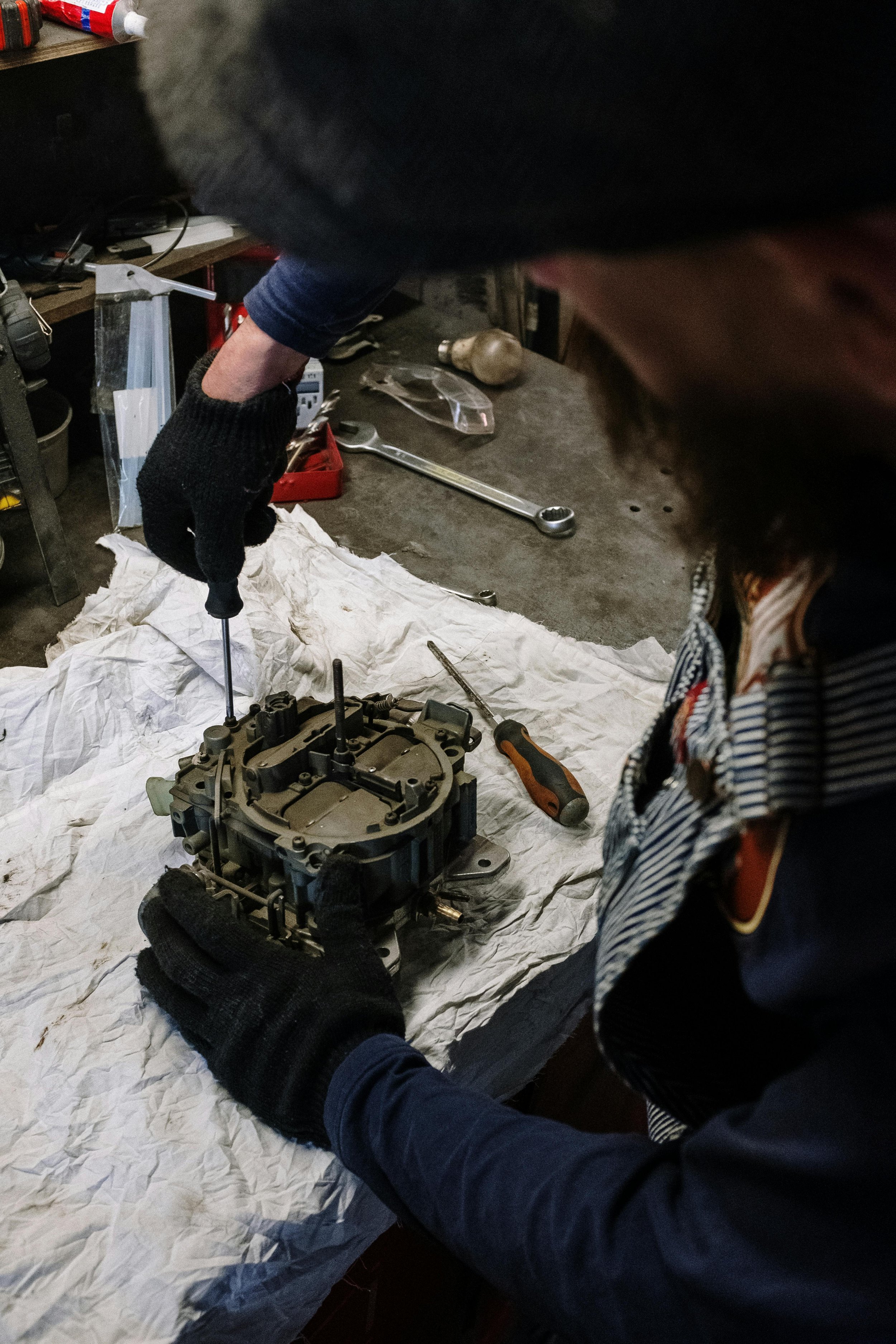 Person working on a mechanical part, possibly an engine carburetor, on a wrinkled white cloth with tools and mechanical components around.