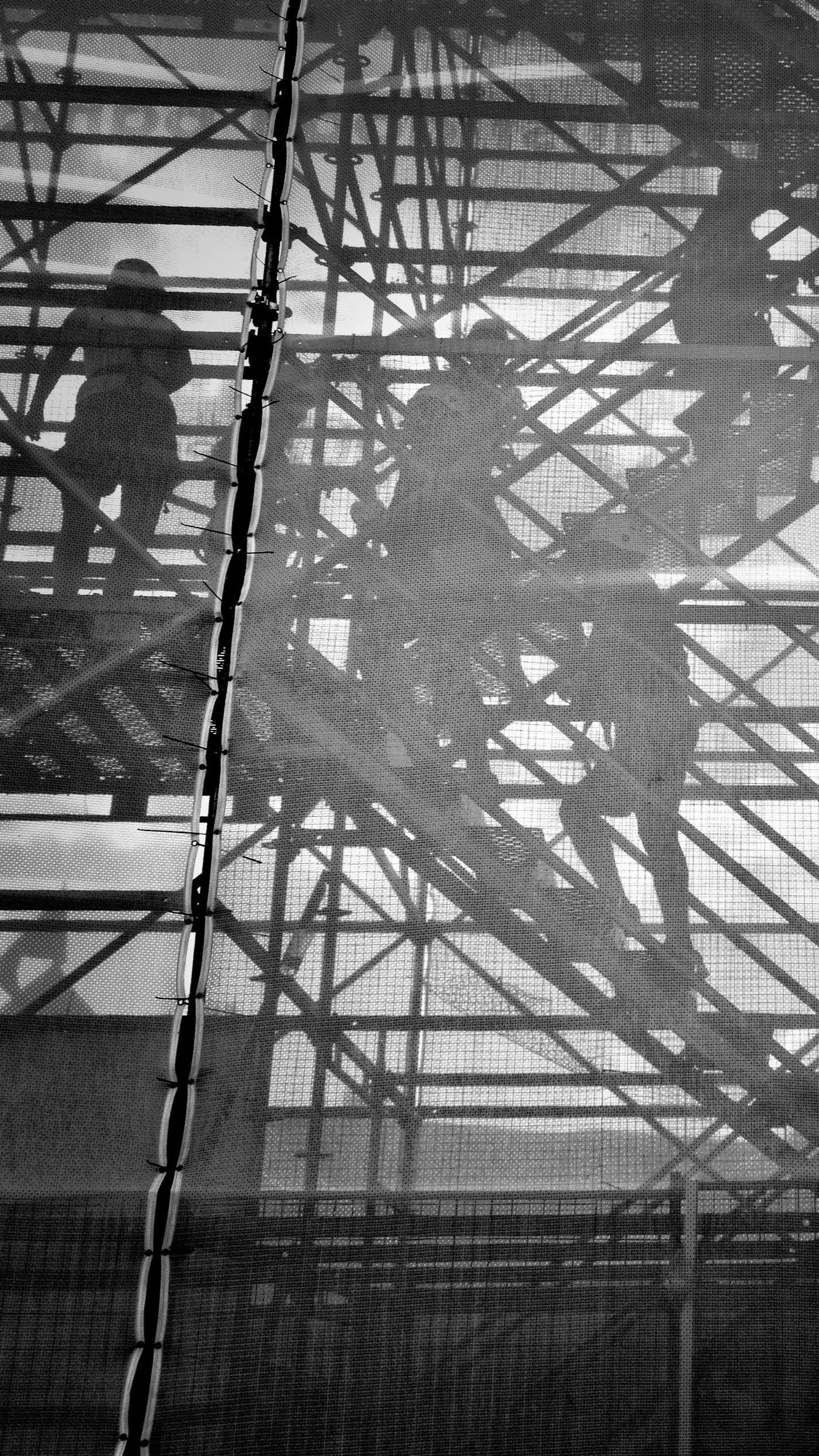 Silhouettes of children playing on a large metal roller coaster structure, viewed through a mesh fence.