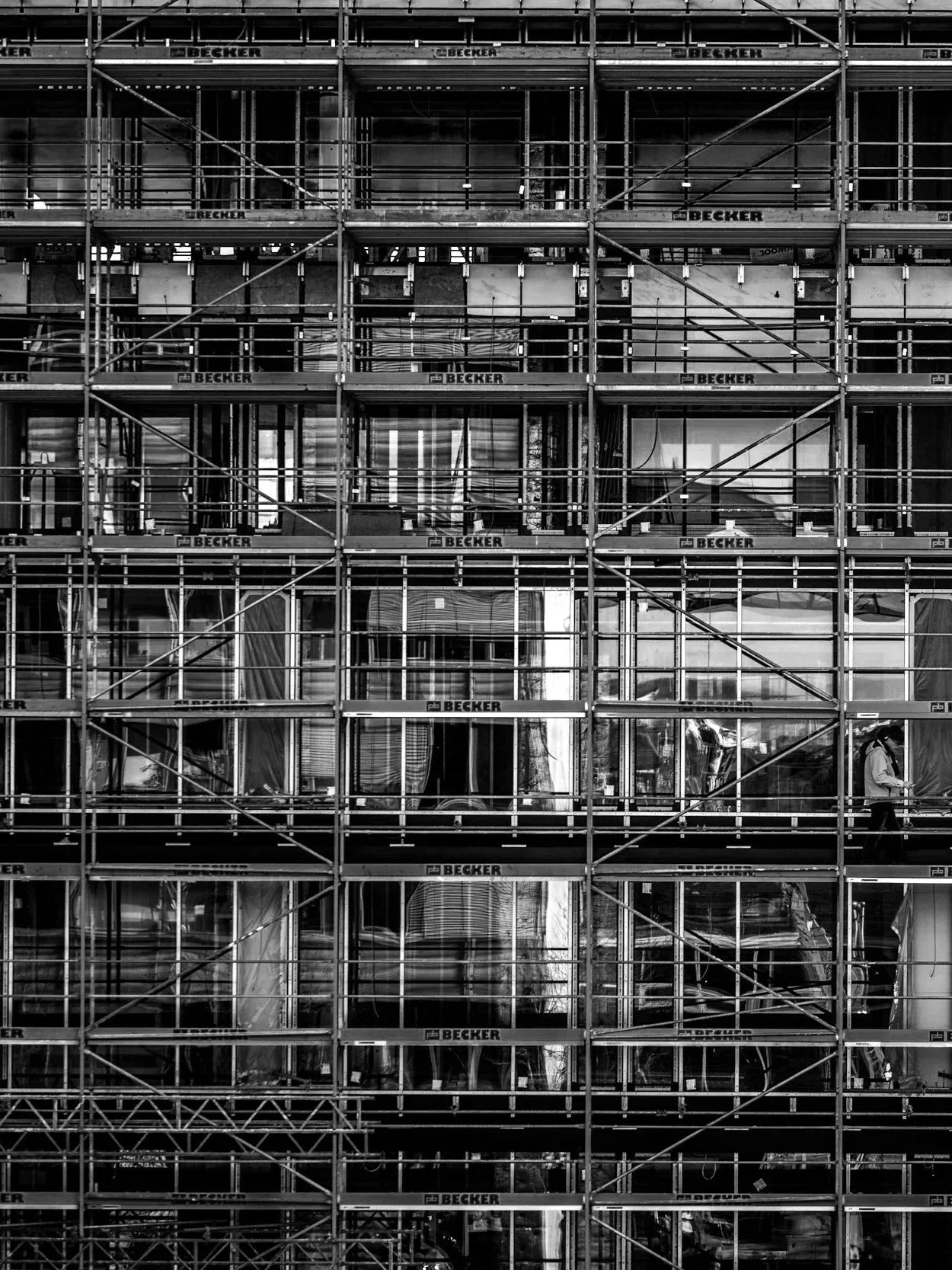 A building under construction covered with scaffolding and protective netting, with some reflective windows showing the surroundings.