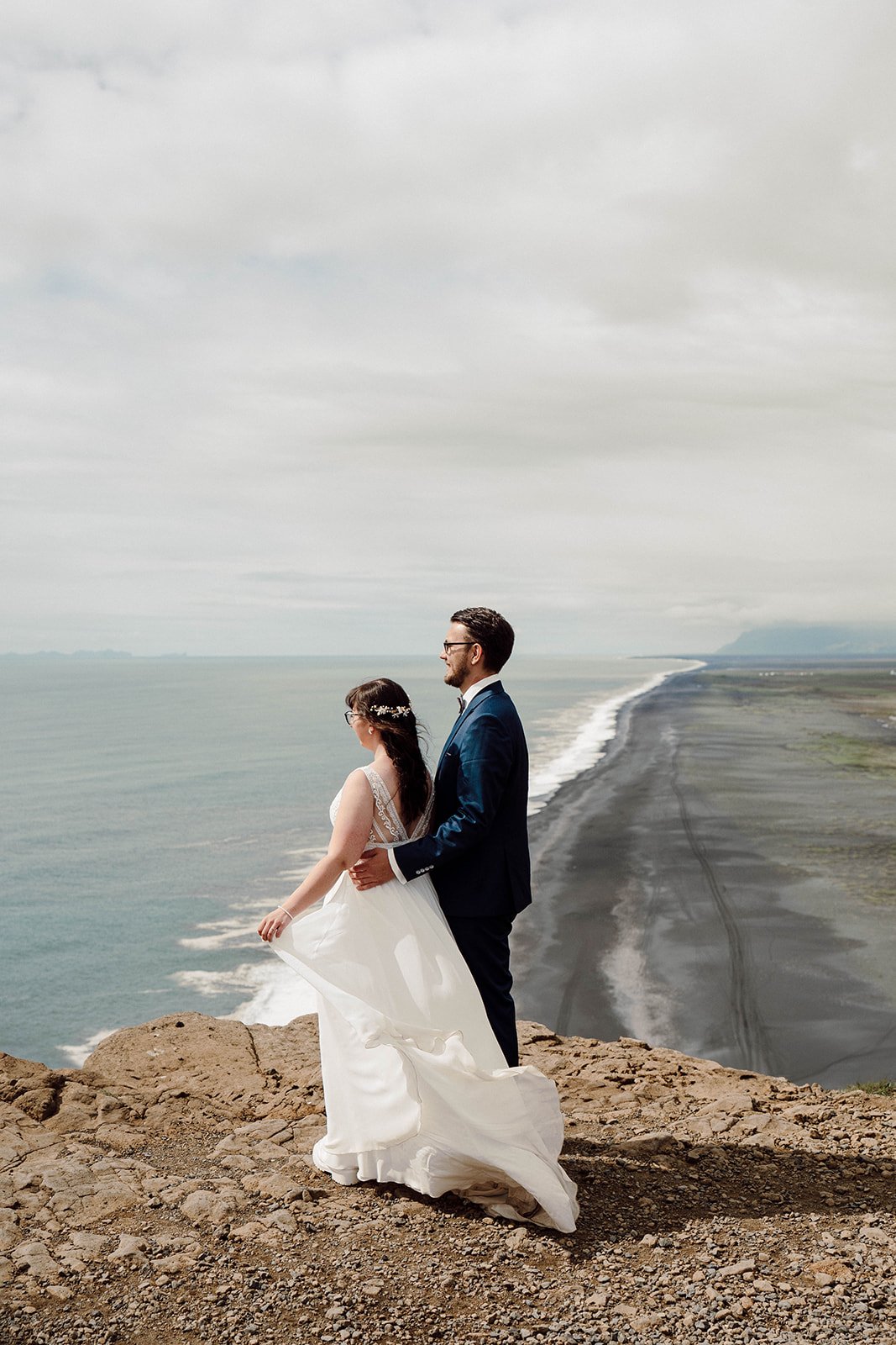 Bride and groom standing on a cliff overlooking Iceland’s black sand beach and ocean