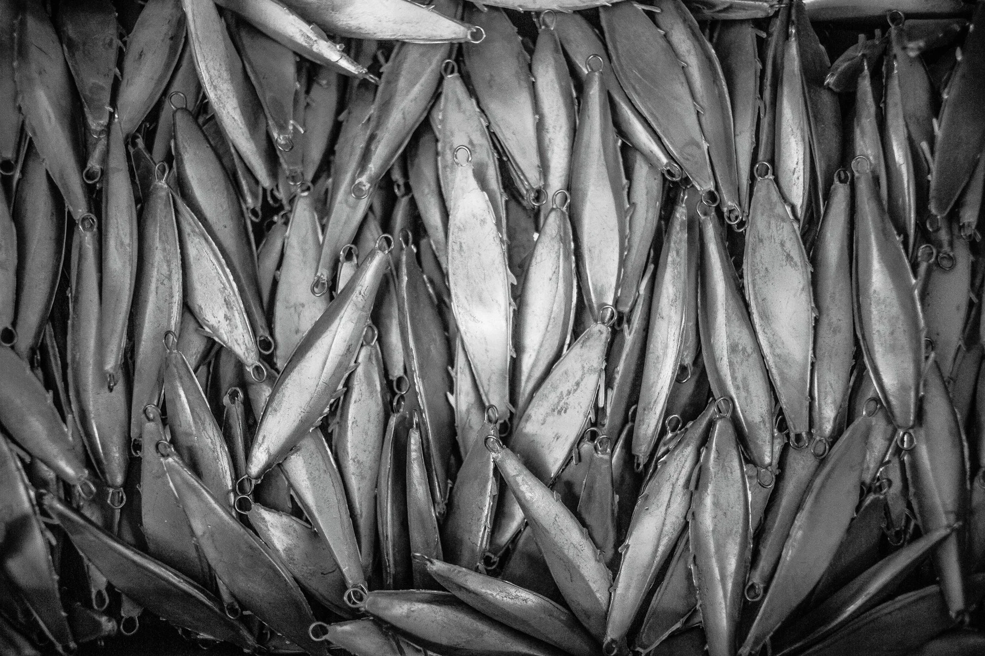 A pile of numerous metal fishing lures with hooks, arranged in a densely packed pattern.