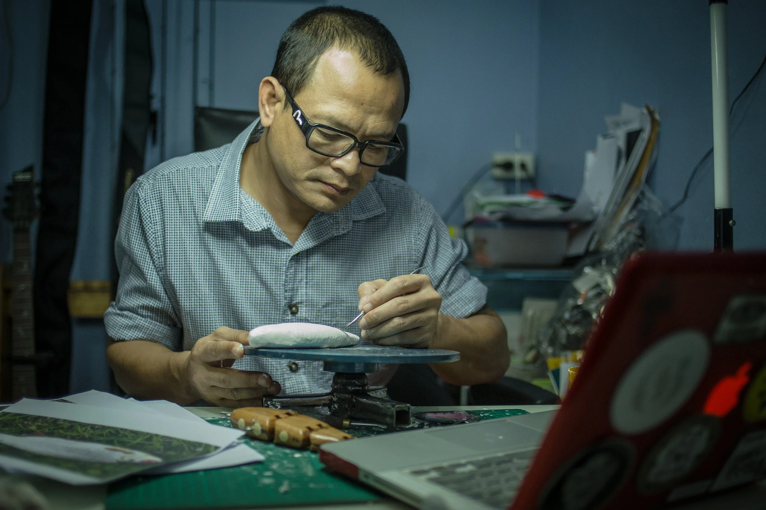 A man wearing glasses and a checkered shirt working on a small object at a cluttered desk with papers, a laptop, and various tools.