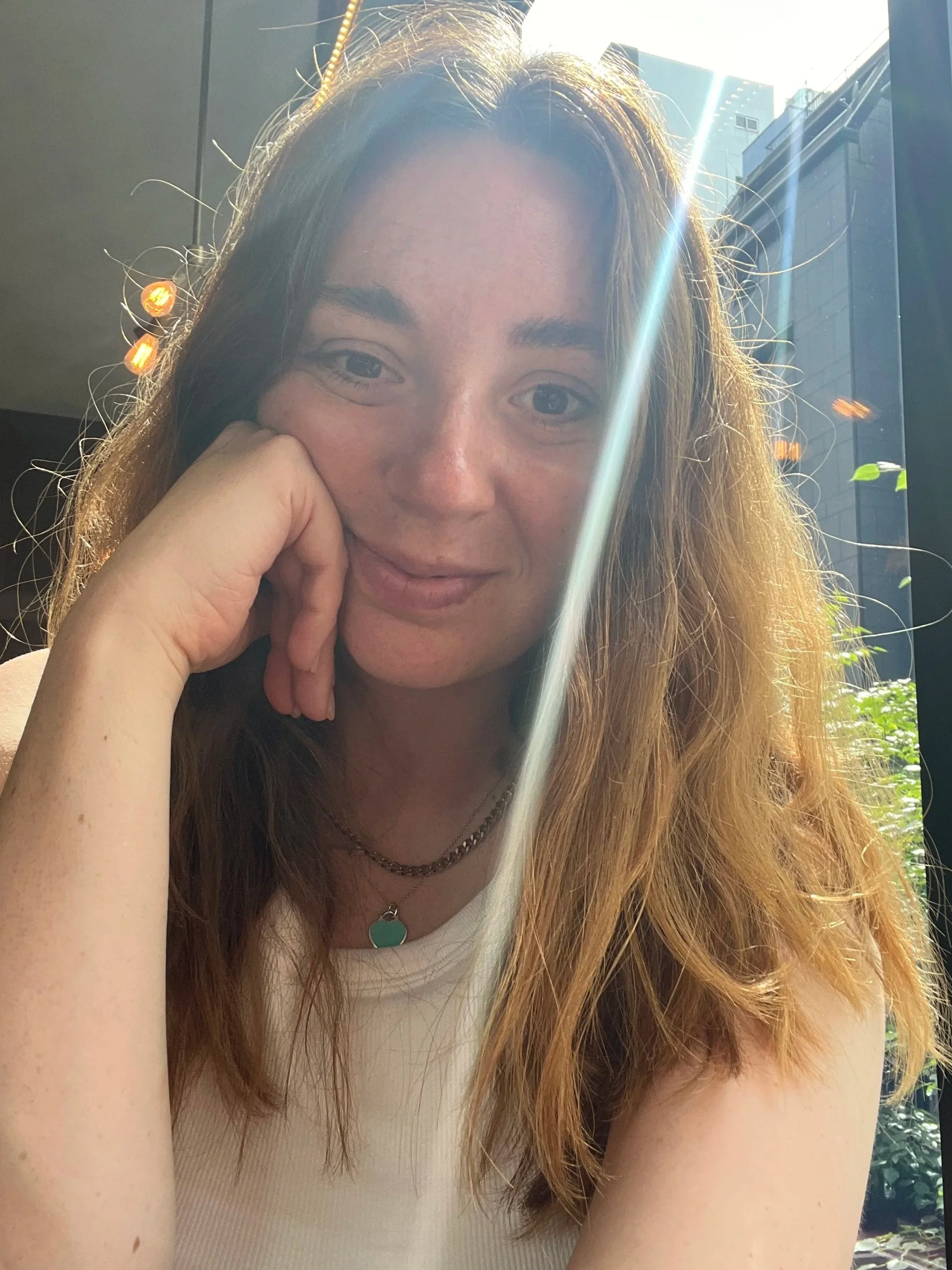 A young woman with long wavy hair and fair skin, smiling gently while resting her face on her hand. She is sitting indoors near a large window with sunlight and buildings visible outside.