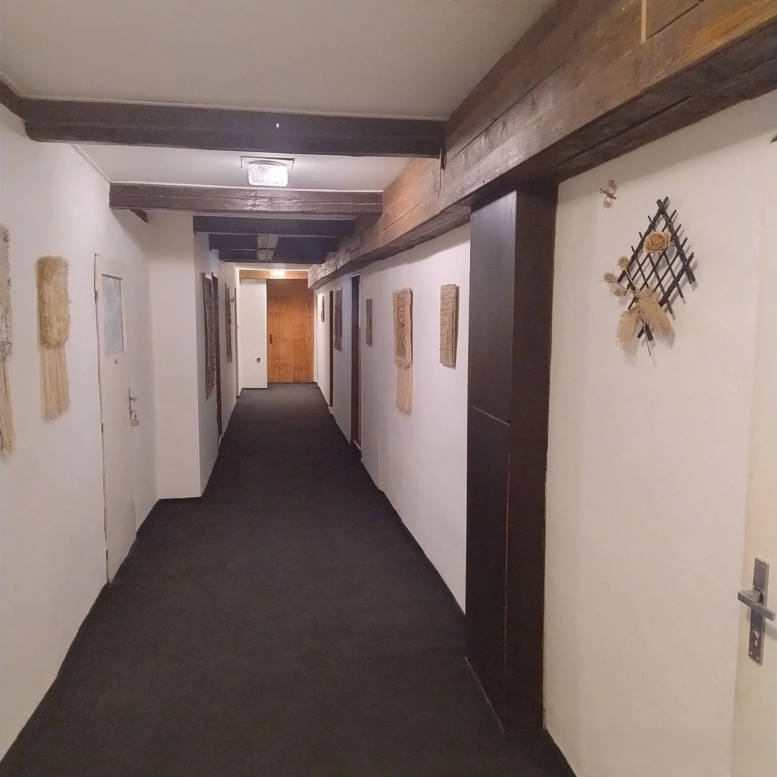 A hallway with white walls, dark wood beams on the ceiling, and various textile art and decorative pieces on the walls. The floor is covered with dark carpet, and a wooden door is visible at the end of the hallway.