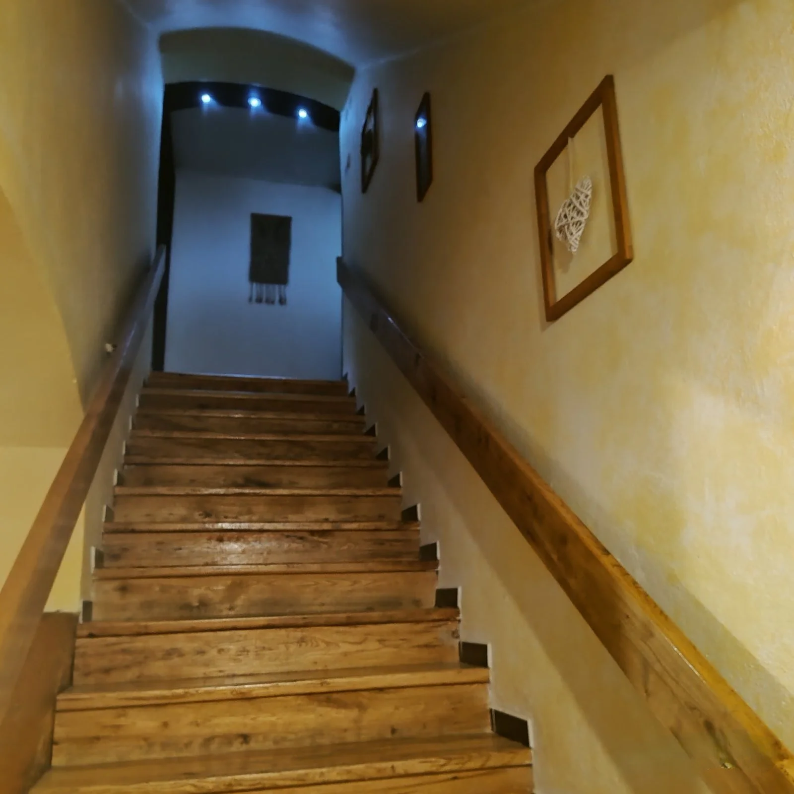 Wooden staircase with yellow walls and framed artwork, leading to a dimly lit corridor with wall hangings and blue ceiling lights.