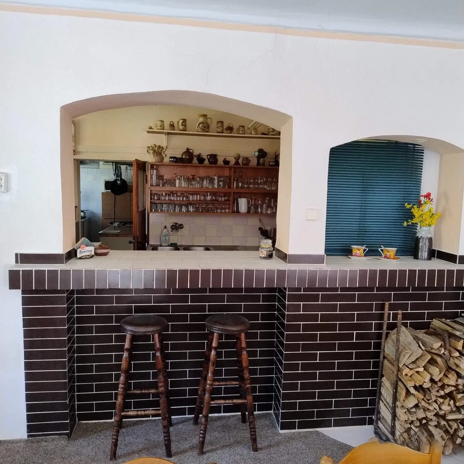 A kitchen viewed through a serving window with a dark tiled counter and two wooden bar stools in front. Inside the kitchen, walls are lined with shelves holding jars, pots, and glasses. To the right, a small area with a vase of yellow flowers and oth
