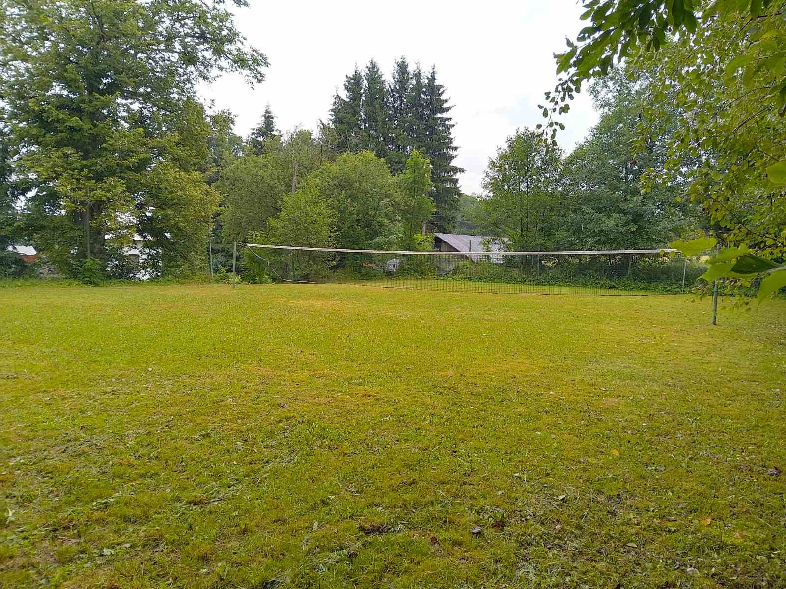 A grassy backyard with a volleyball net stretched across the middle, surrounded by trees and a house in the distance overcast sky.