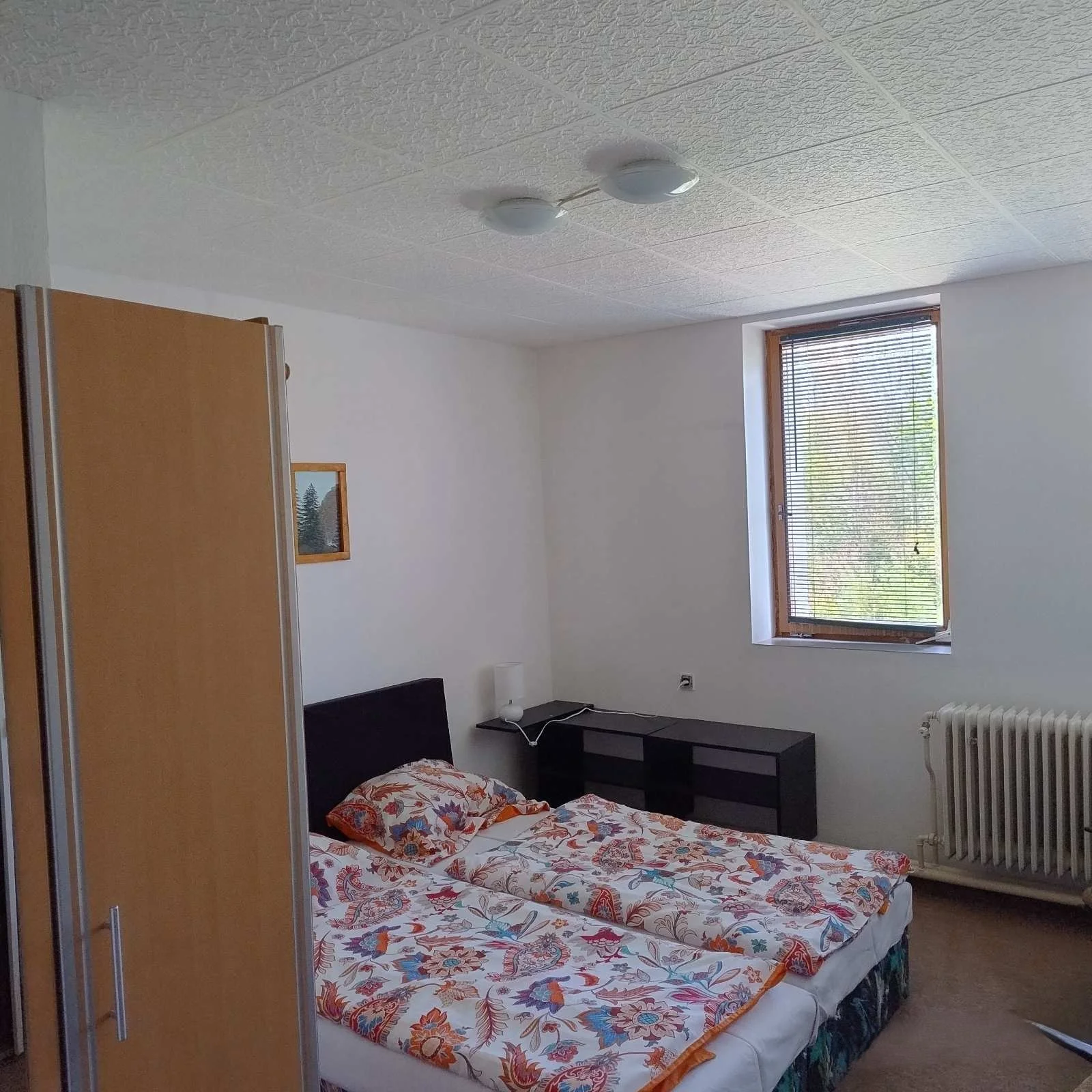 A simple bedroom with a bed covered in a floral patterned bedspread, a black headboard, a small black nightstand with a lamp, a window with blinds, a radiator, and a wooden wardrobe.