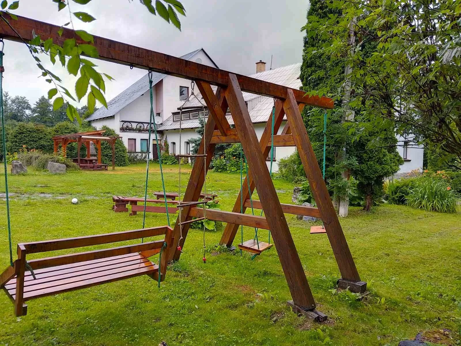 A wooden swing set with two swings and a slide on a grassy yard with shrubs and trees, and a house in the background.