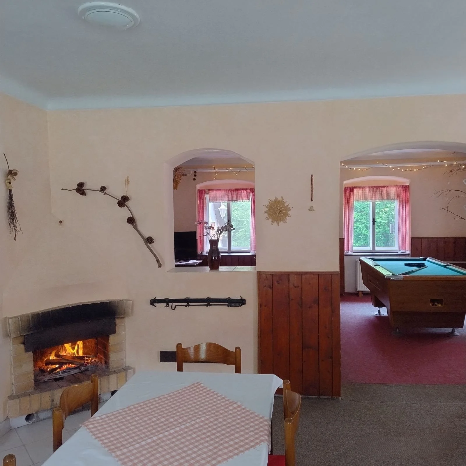 A cozy living room with a fireplace, wooden dining table, and chairs, and a view into a game room with a pool table, decorated with fairy lights and pink curtains, with greenery visible outside the windows.