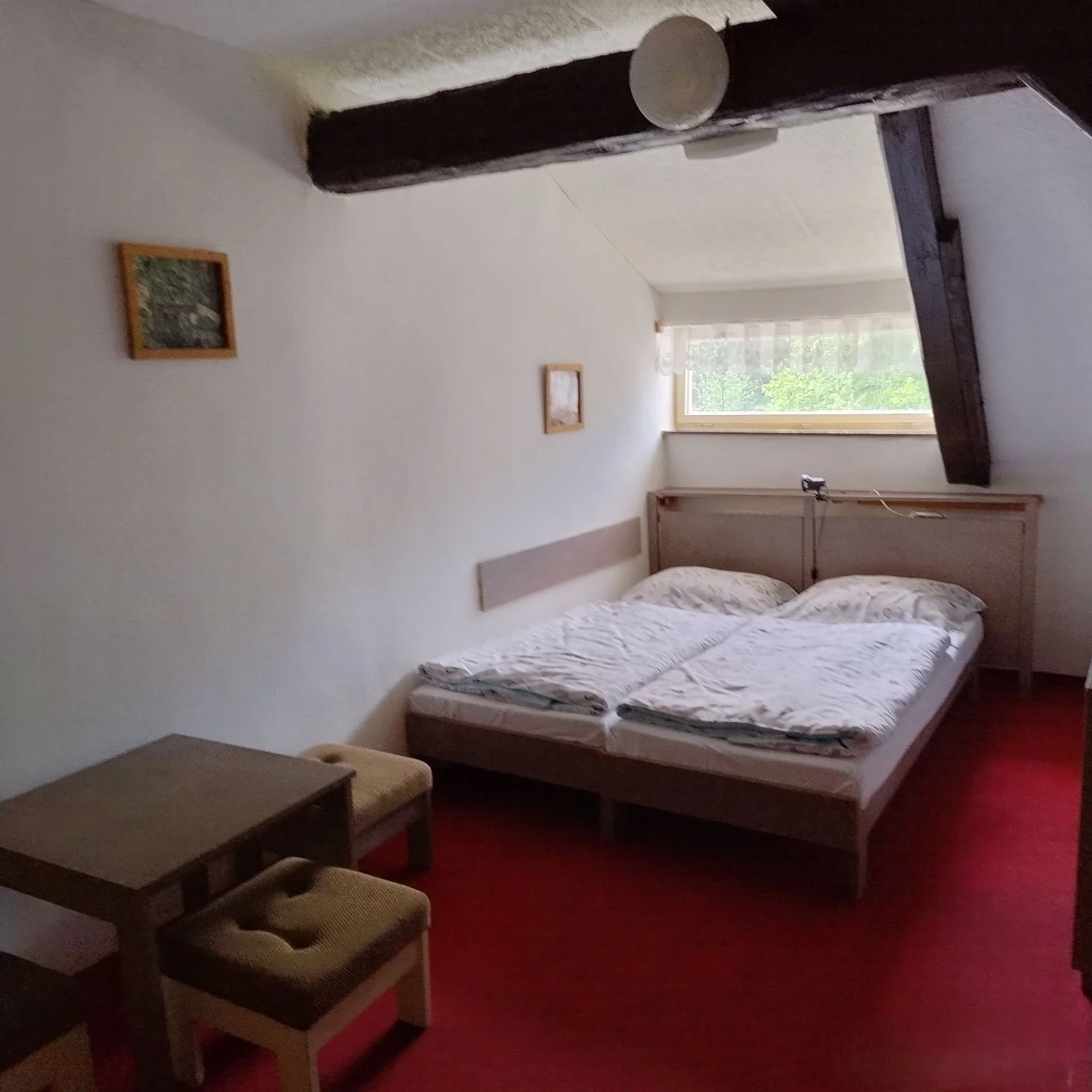 A bedroom with a bed, two pillows, and a white blanket, a small table and two stools on a red carpet, a window with a view of green trees, a wooden beam on the ceiling, and framed pictures on the wall.