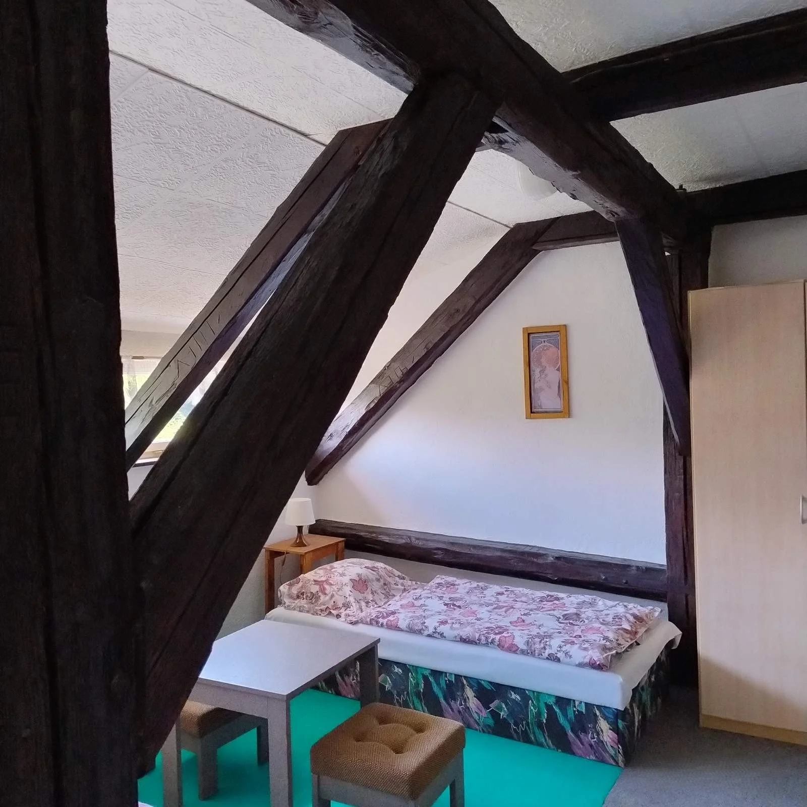 A cozy bedroom with a single bed that has floral bedding, a small nightstand with a lamp, a framed picture on the wall, a blue rug, a wooden stool, and a wardrobe, all under a slanted ceiling with dark wooden beams.